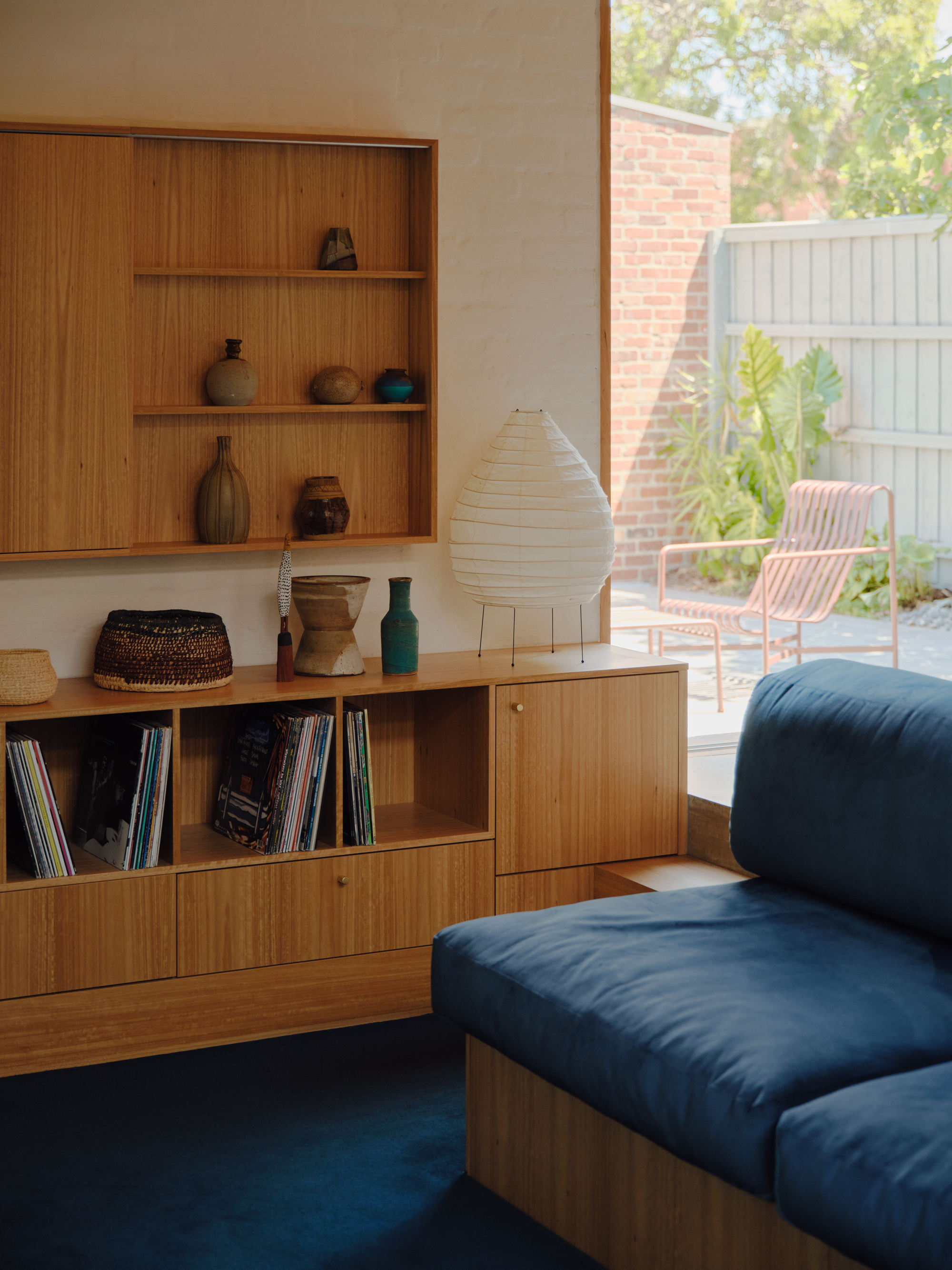 Timber shelving unit with ceramics and records beside glass door overlooking garden with pink chair