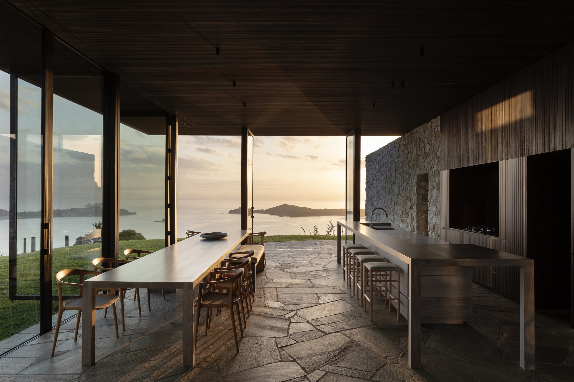 Dining and kitchen pavilion with irregular stone flooring opening to coastal views at sunset