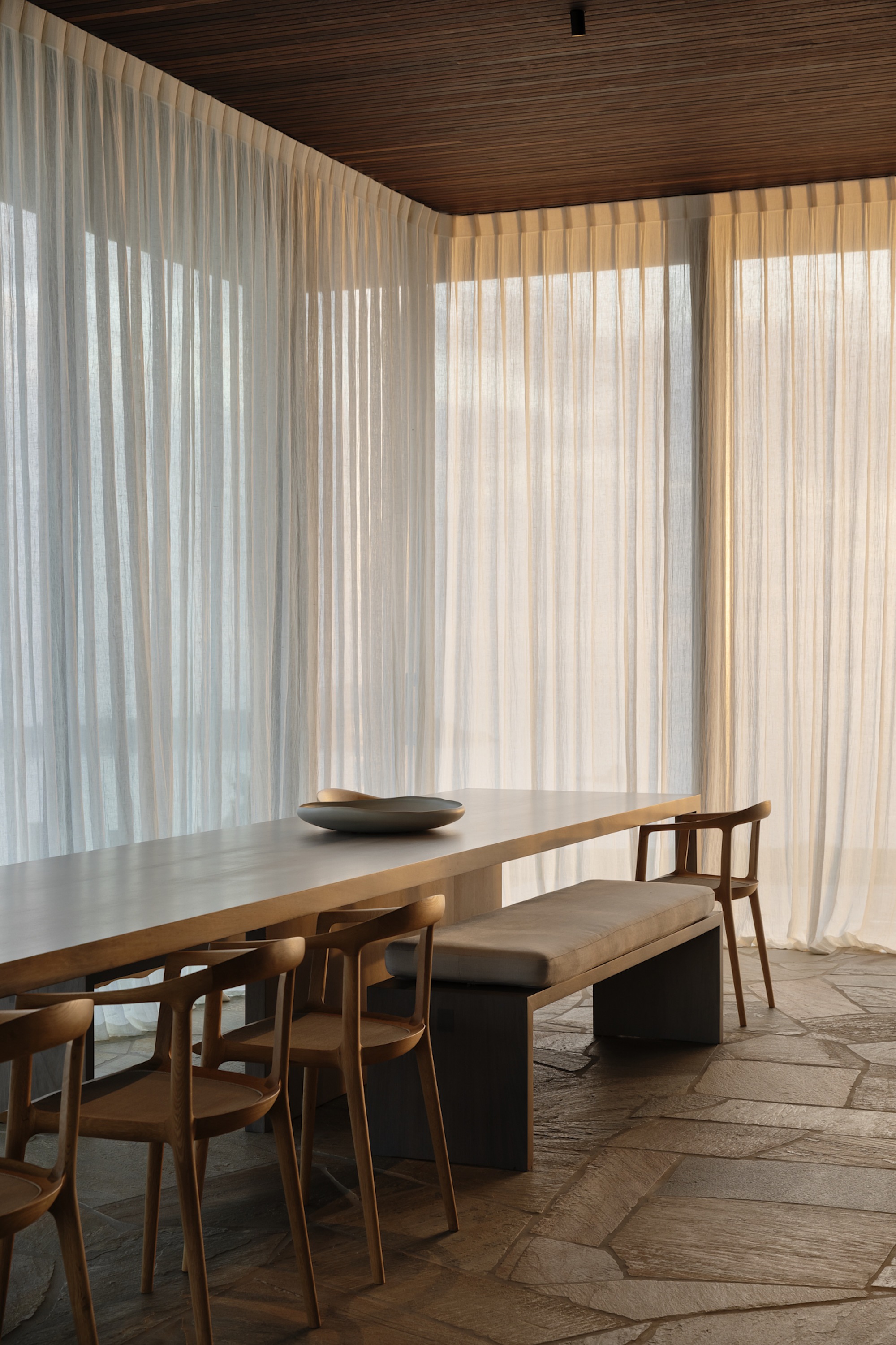 Dining room with timber table and irregular stone floor tiles beneath floor-to-ceiling sheer curtains