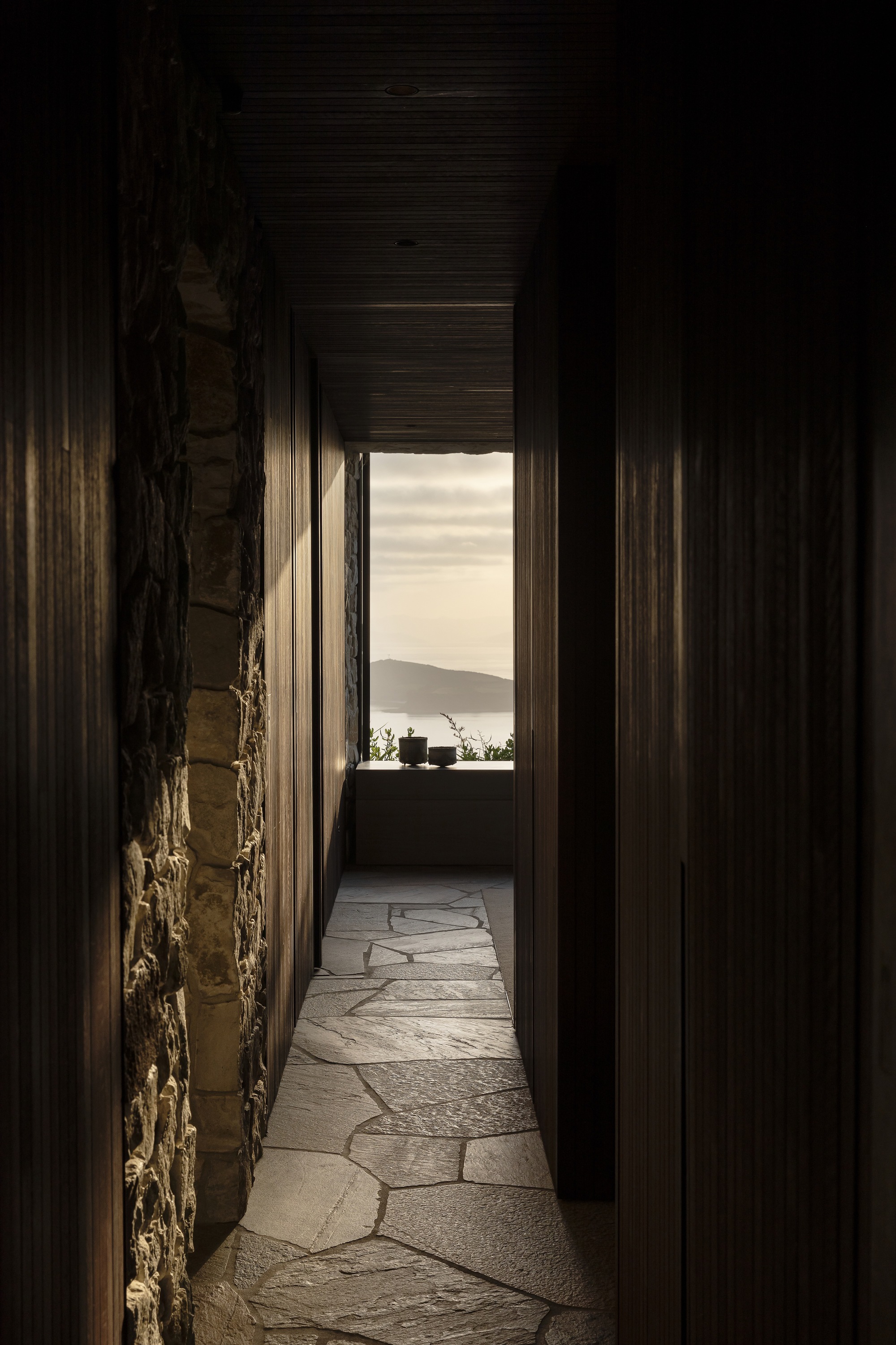 Narrow corridor with dark timber walls and irregular stone flooring leading to distant coastal view