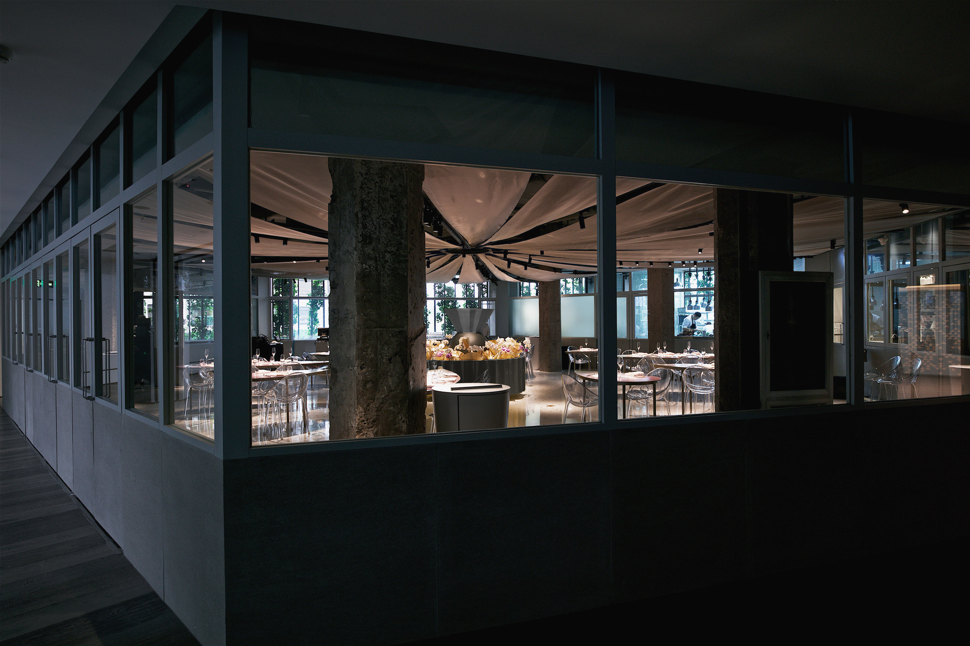 Exterior view through glazed facade showing the illuminated dining room with draped ceiling at dusk