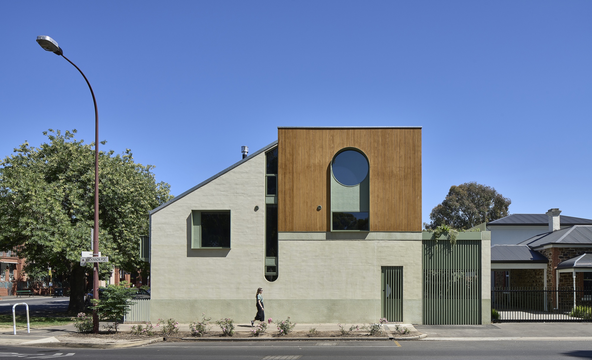 Street facade with vertical timber cladding, round window, and pale render base with pedestrian passing
