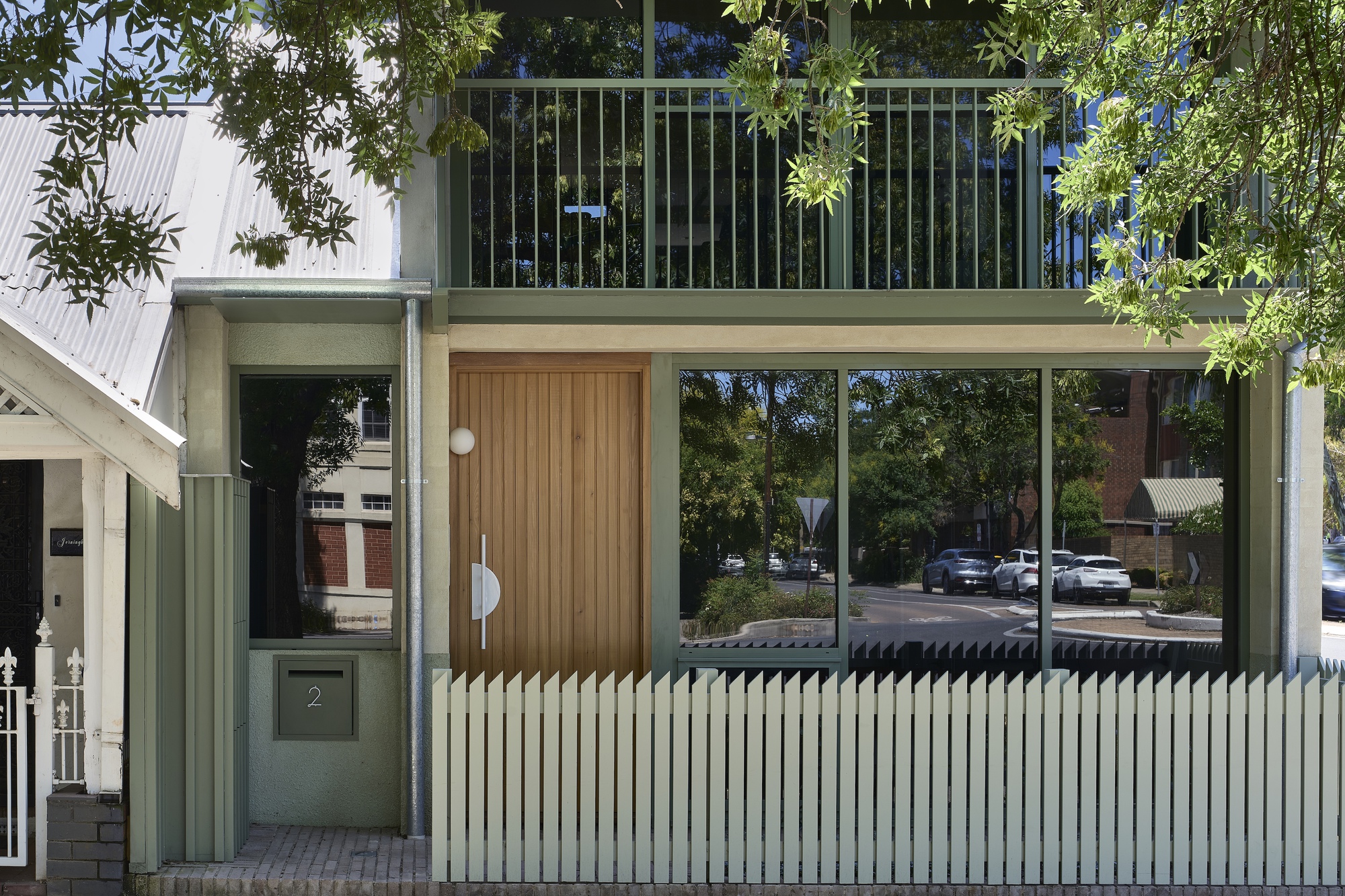 Ground floor entry with vertical corrugated metal fence, timber door, and glazed balcony above among trees