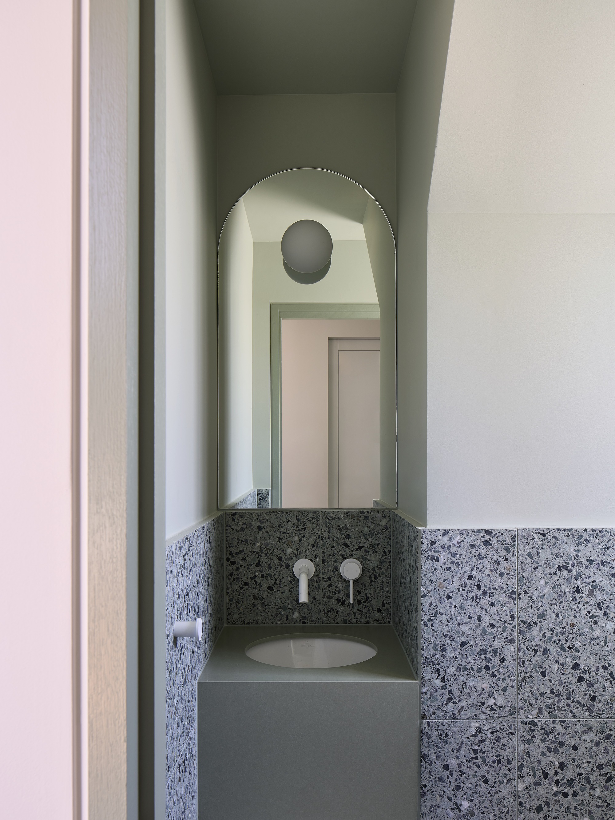 Powder room vanity niche with terrazzo wainscot, round mirror, and arched opening to hallway beyond