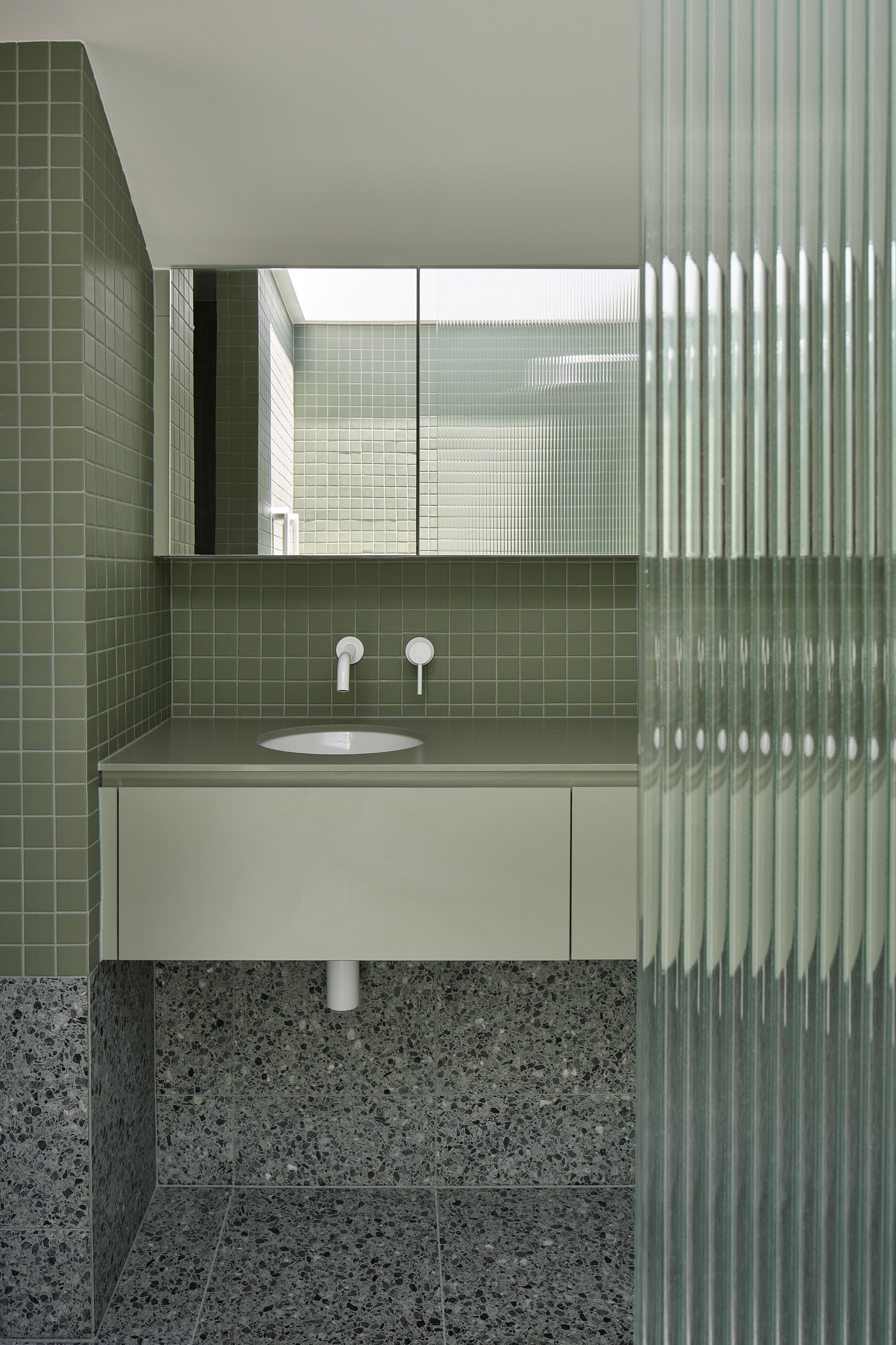Bathroom with olive green square tile walls, terrazzo flooring, and vertical fluted glass screen beside floating vanity