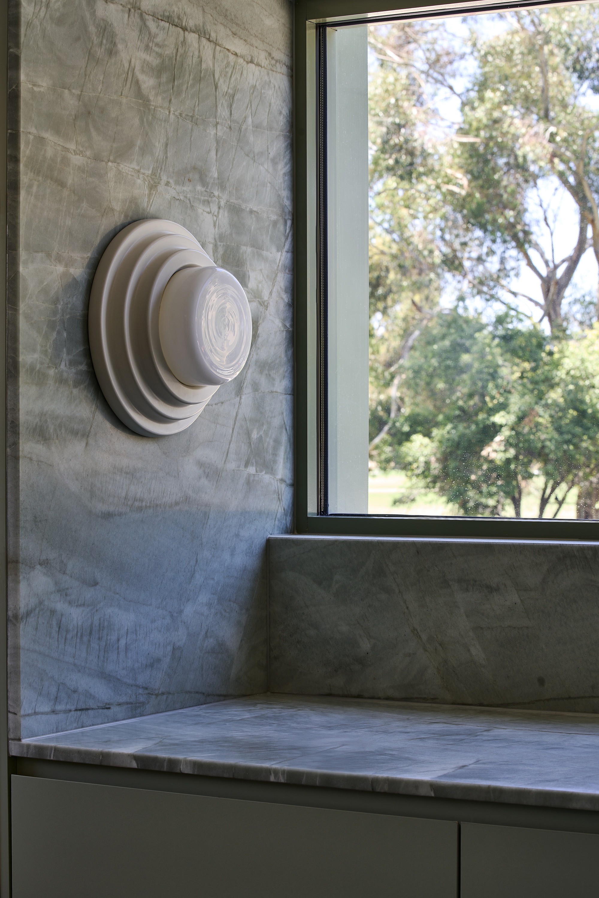 Circular wall-mounted light fixture on concrete wall beside a window framing leafy trees outside