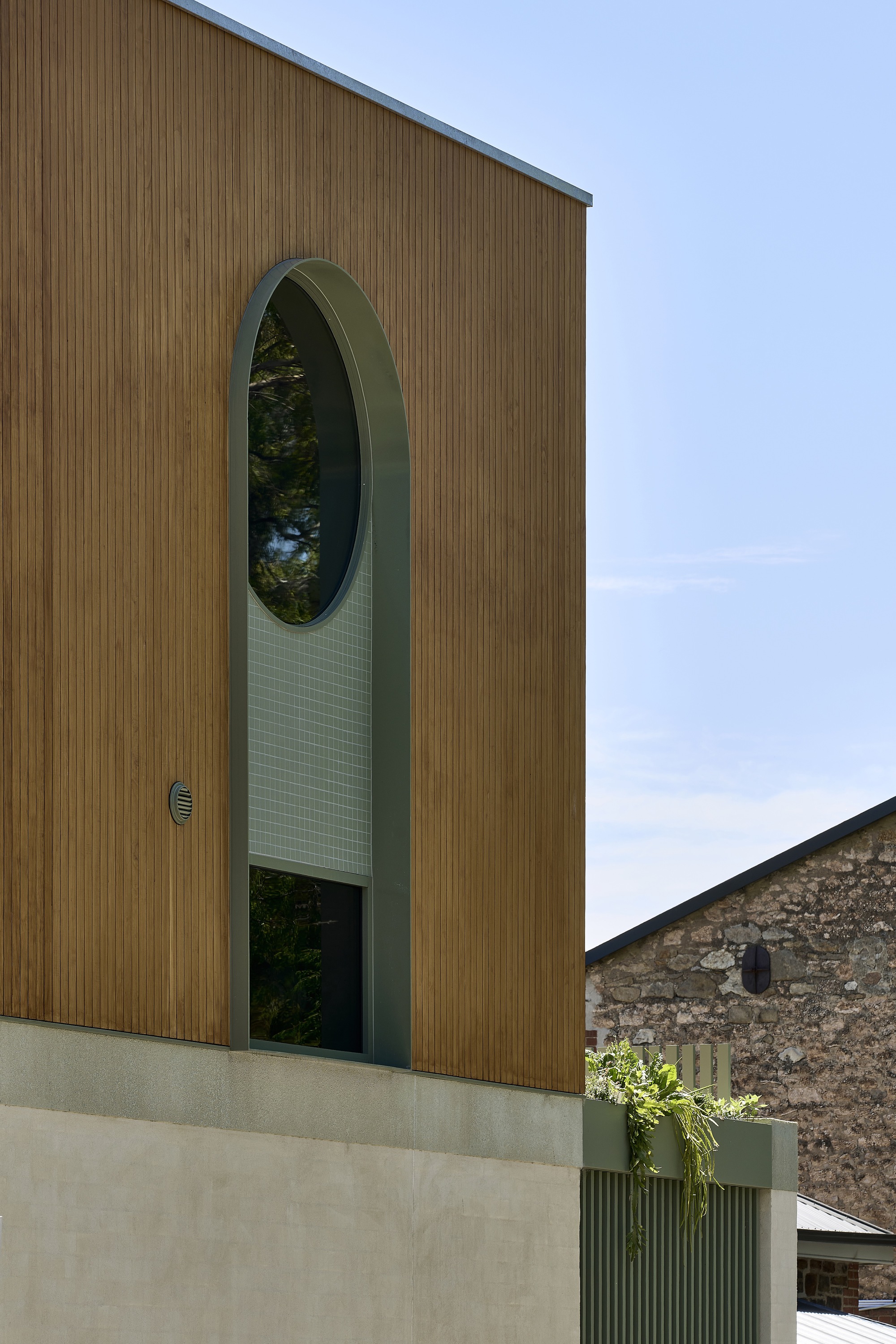 Vertical timber cladding facade with tall arched window opening and tiled green surround under blue sky