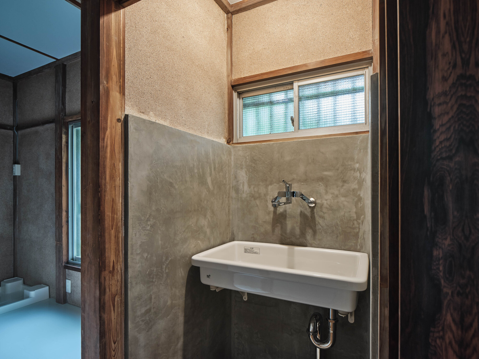 Bathroom corner with wall-mounted sink beneath a small window and textured plaster walls framed in timber
