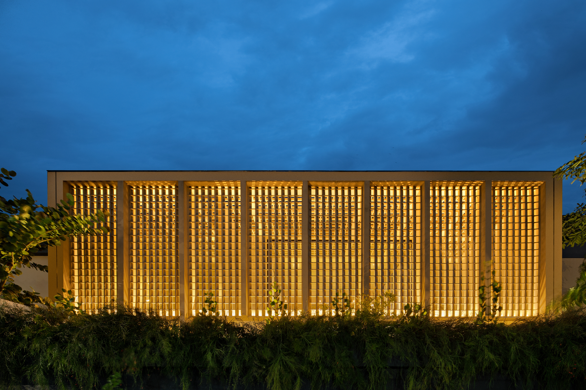 Illuminated perforated brick screen facade glowing against blue evening sky with landscaped foreground