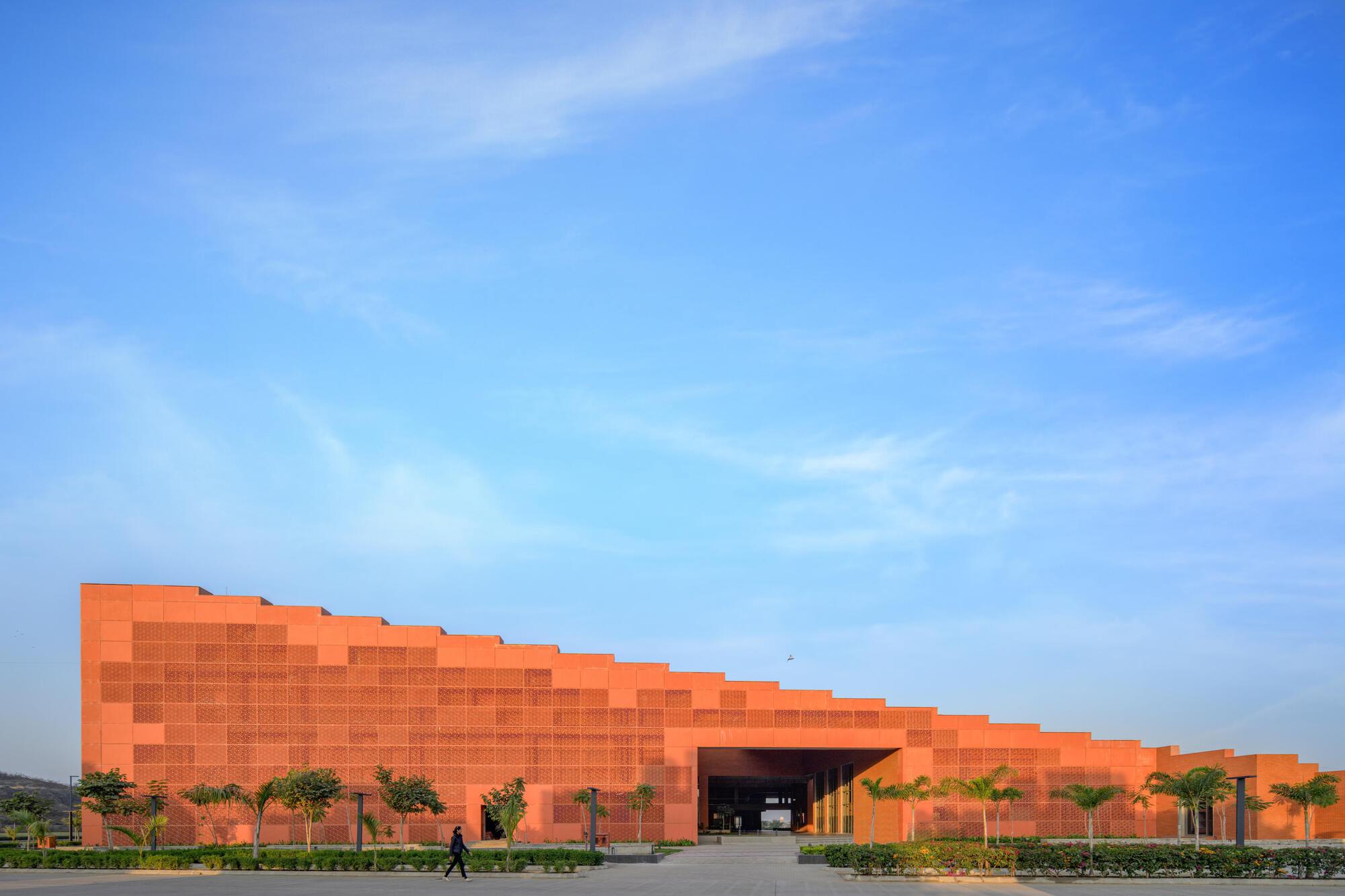 Long facade of pixelated orange brick panels with a central entrance flanked by palm trees