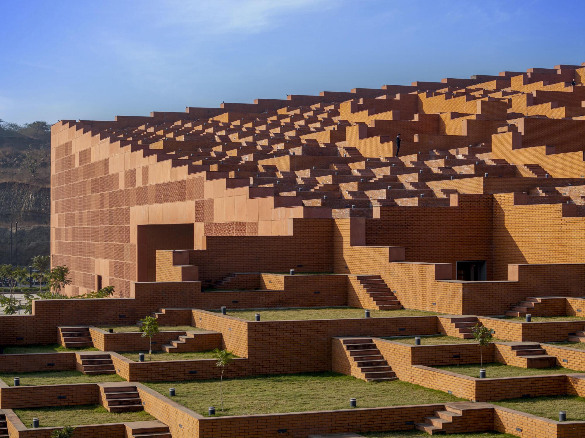 Cascading brick facade with pixelated massing of orange and brown blocks beneath a clear blue sky