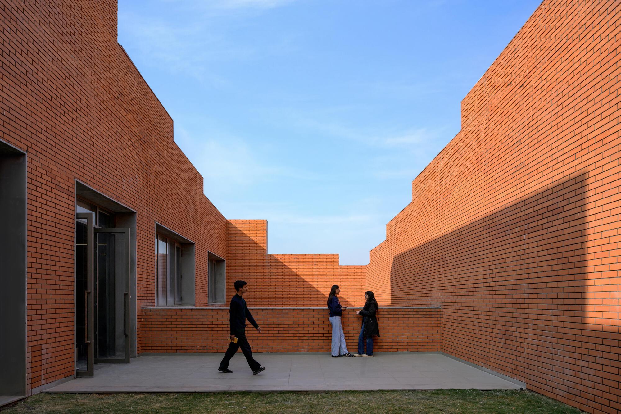 Courtyard enclosed by stepped brick walls with three figures walking in late afternoon light