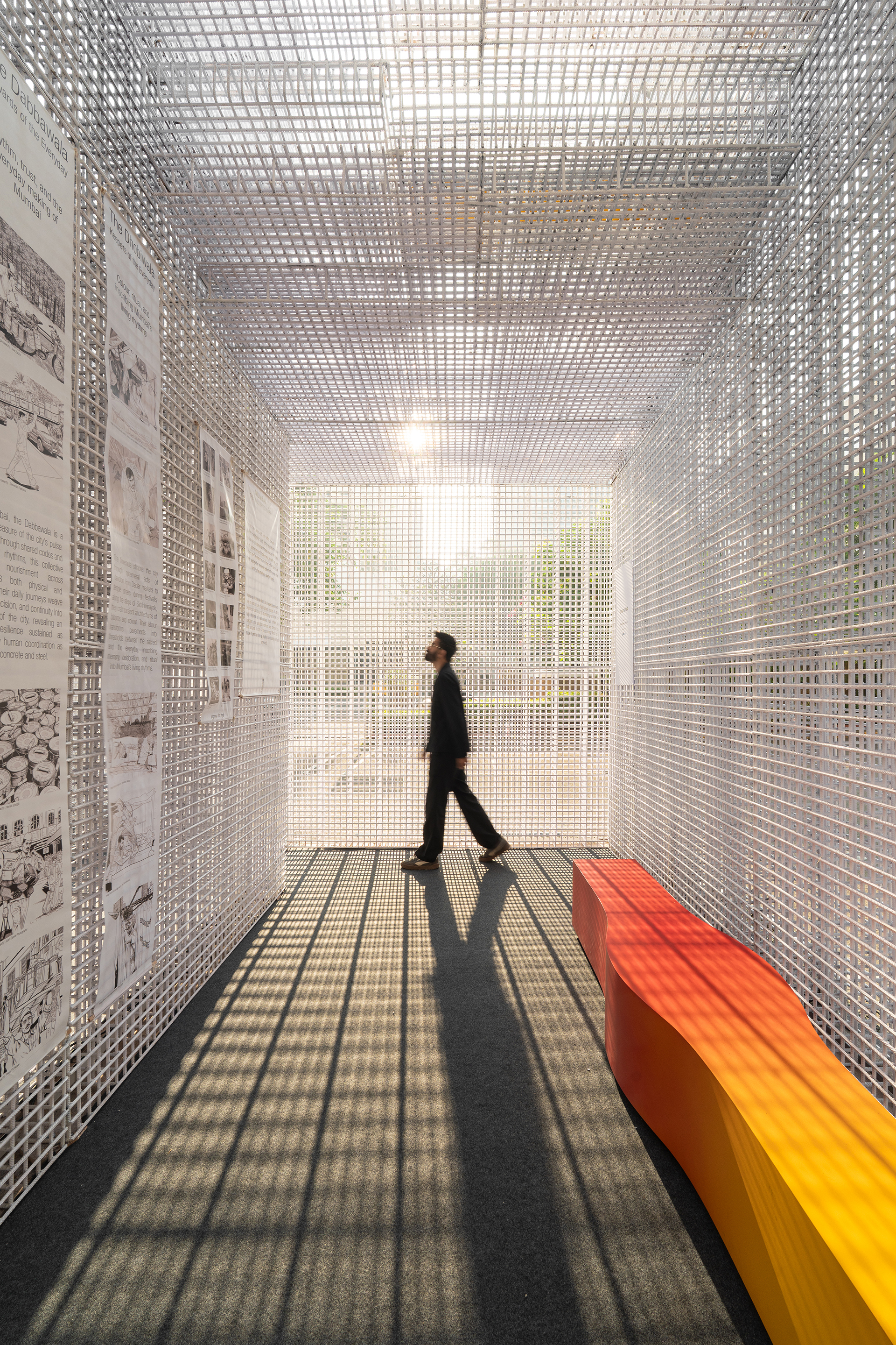 Gallery corridor with perforated white mesh walls and ceiling as figure walks past gradient orange bench