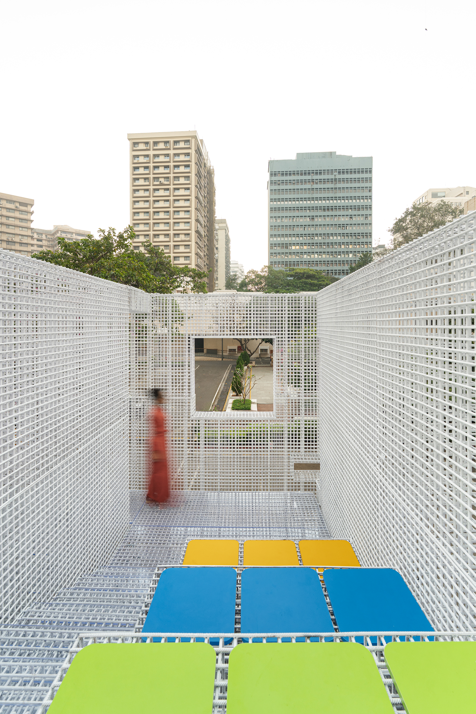 White perforated metal screen corridor with colored floor panels and a visitor in red framed by urban towers