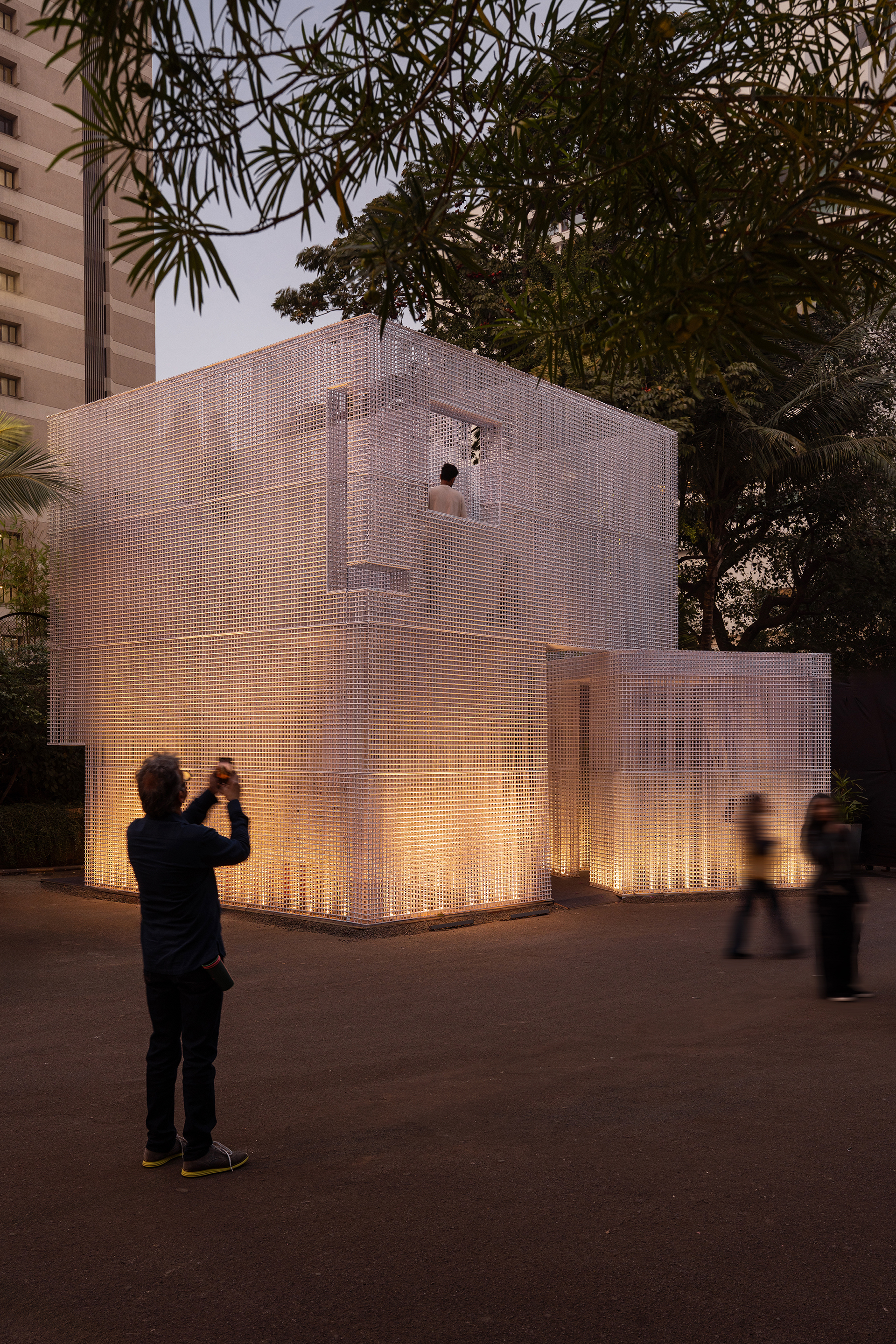 Perforated translucent pavilion glowing from within at dusk as visitors photograph the facade