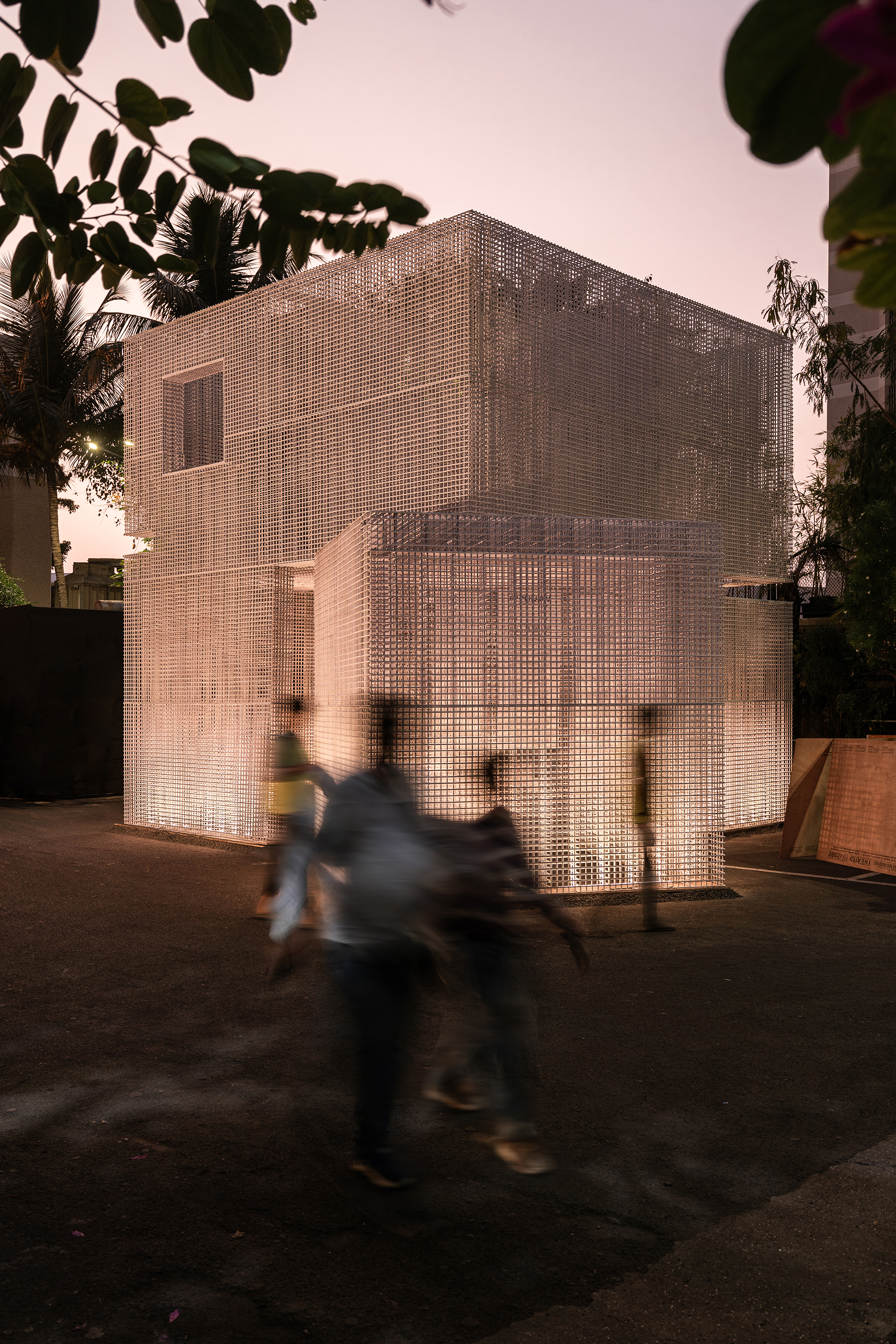 Two-story pavilion wrapped in translucent perforated screens with a motorbike passing at dusk
