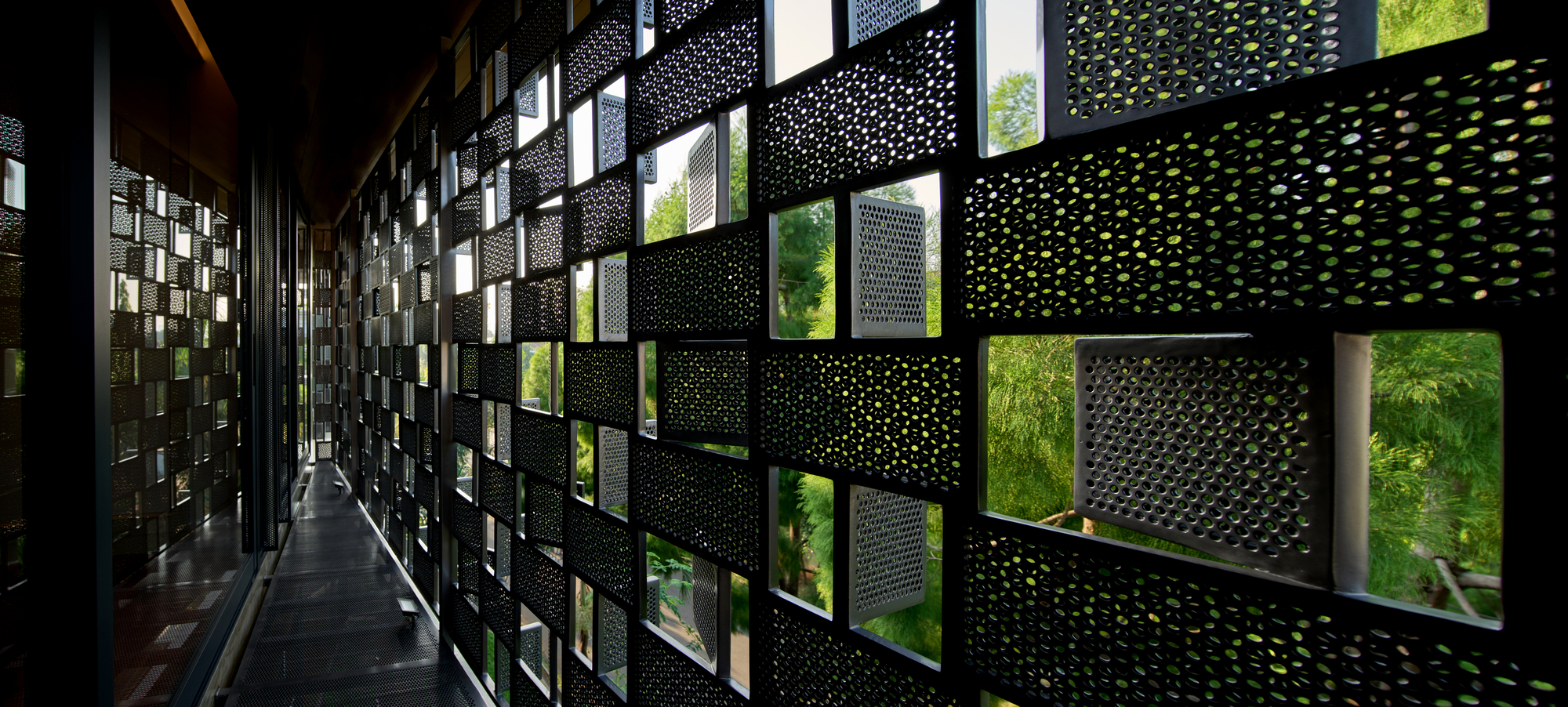 Interior corridor with perforated metal screen wall casting dappled shadows and framing green foliage outside