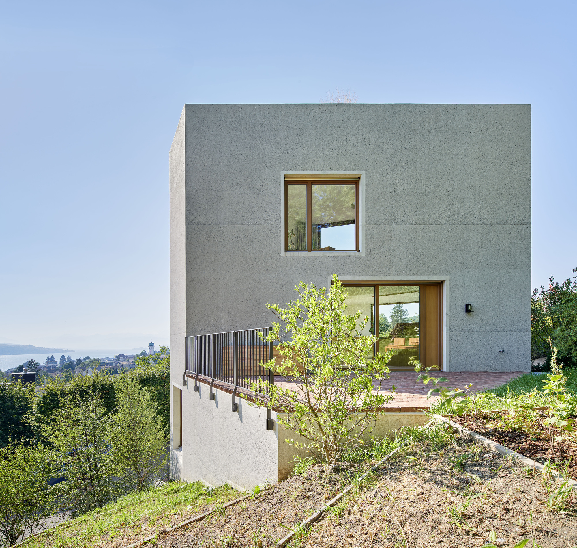 Rear elevation with timber balcony screen and young tree on sloped garden overlooking distant lake