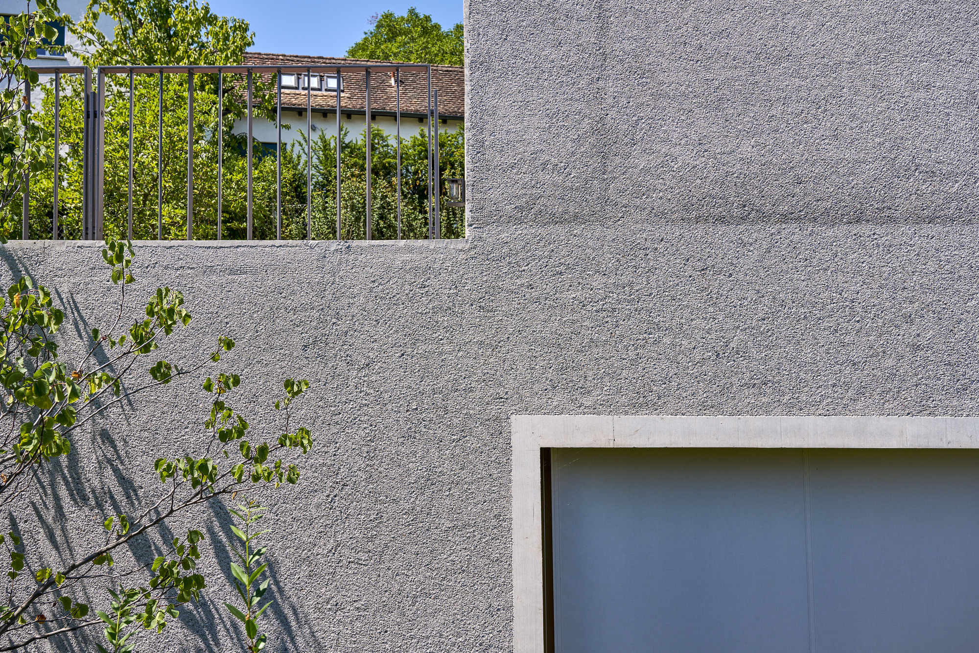 Close-up of textured concrete wall panels with metal railing and young tree casting shadows in sunlight