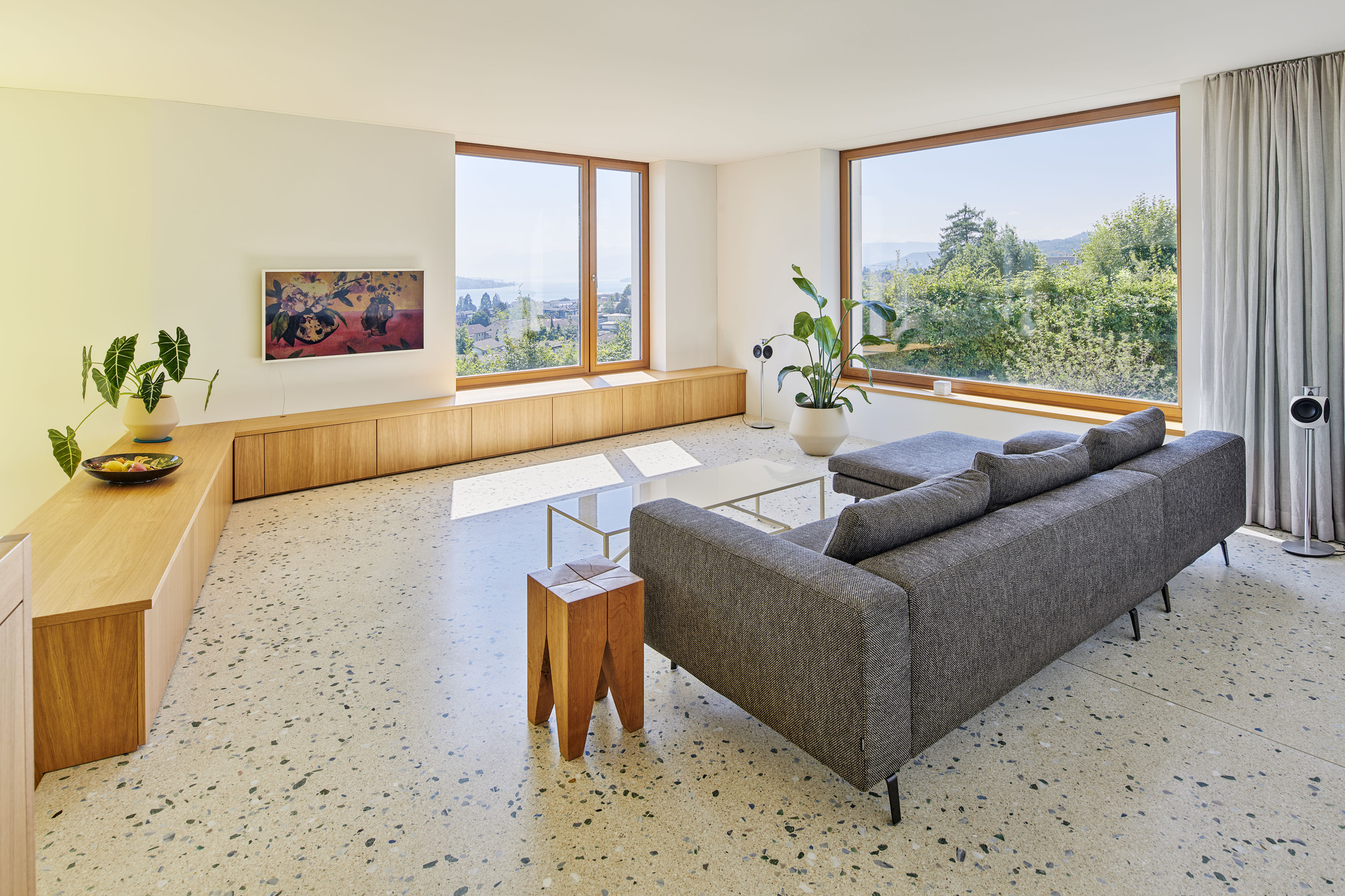 Living room with terrazzo flooring and timber-framed windows overlooking treetops in afternoon light