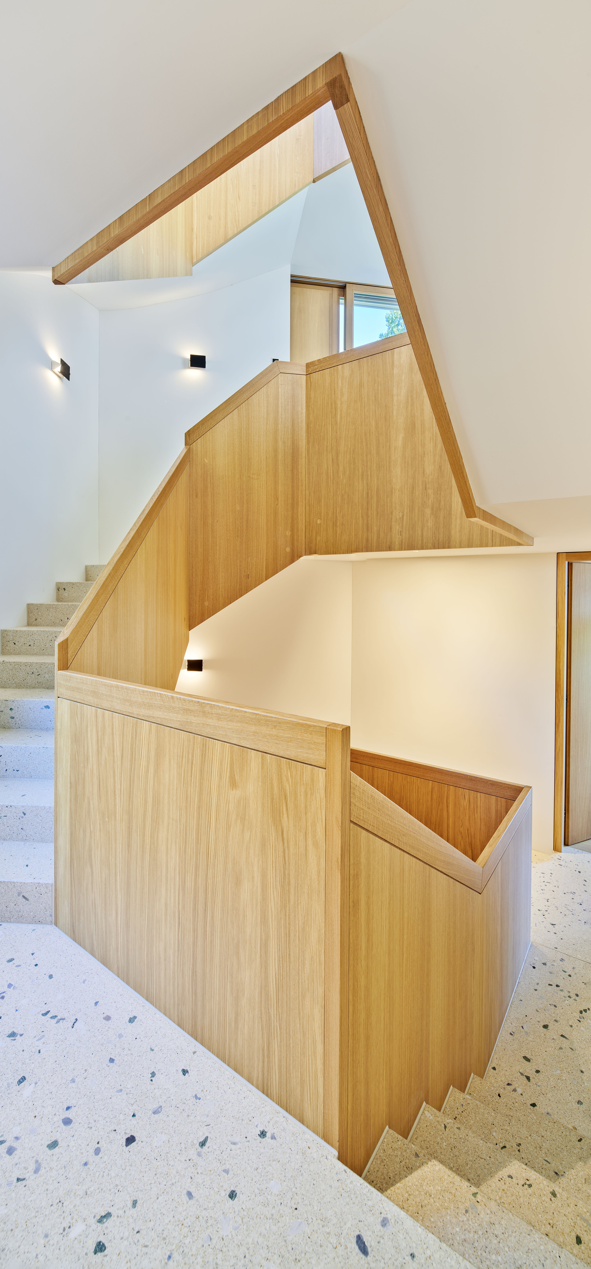 Angular timber-clad stair volume with terrazzo treads connecting multiple levels under a skylight
