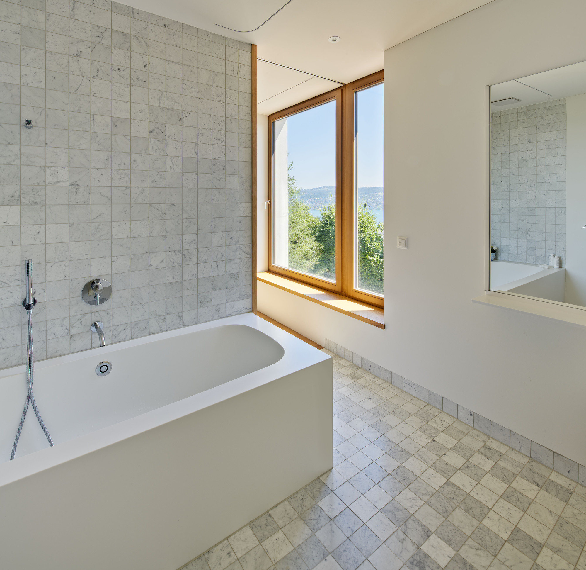 Freestanding tub beneath corner window with pale stone tile wall and distant hills visible outside