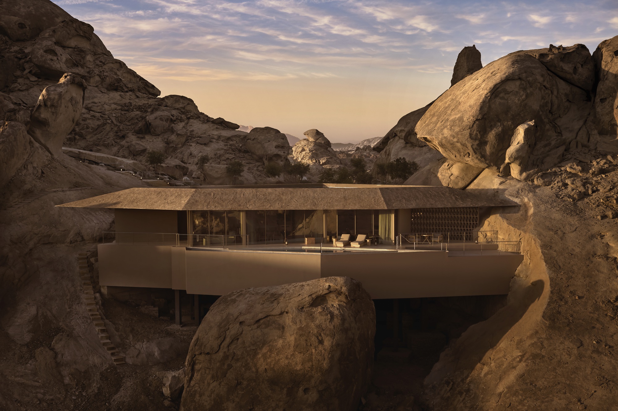 Cantilevered concrete terrace with glass walls framed by massive boulders at sunset