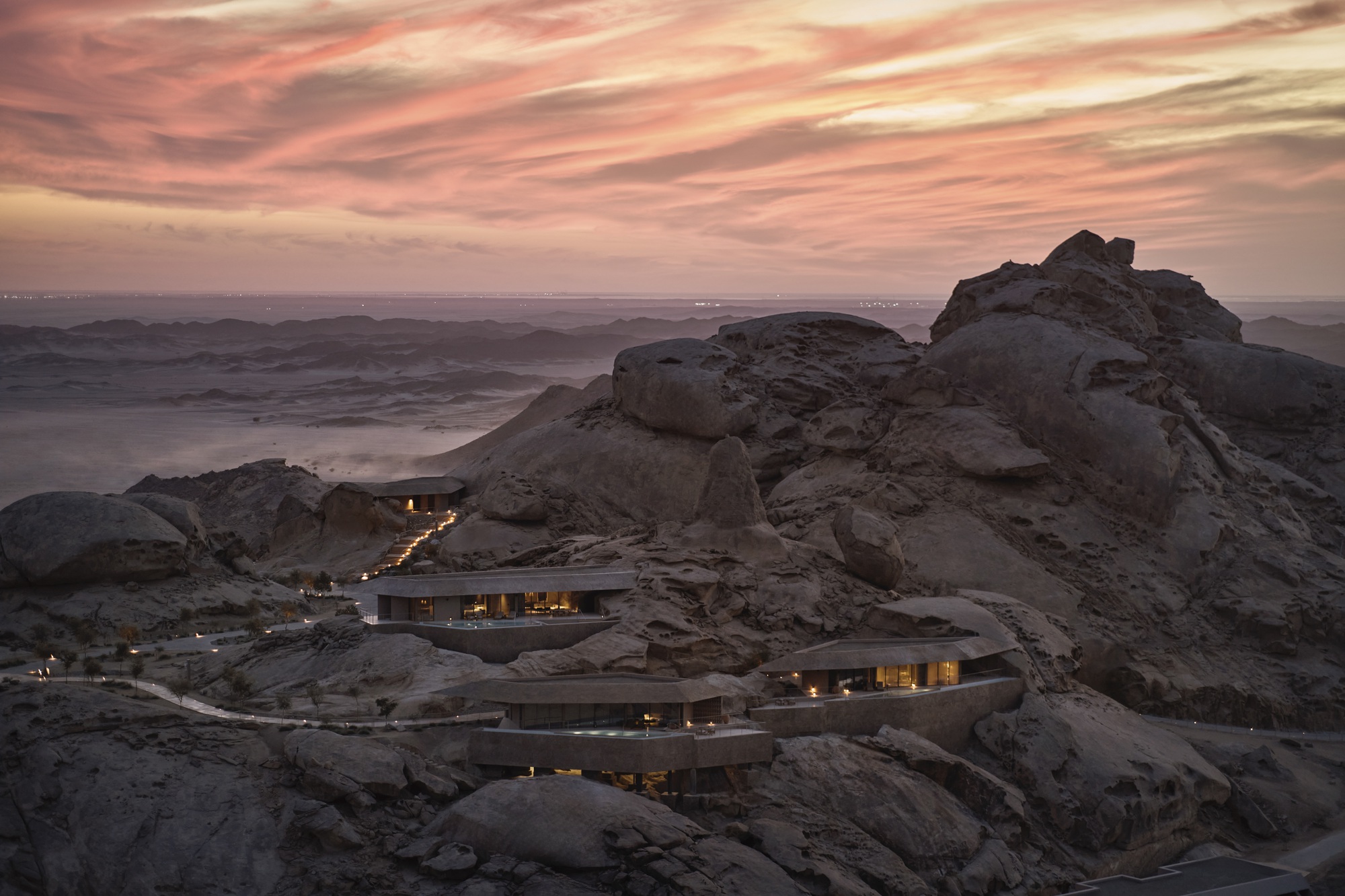 Terraced volumes nestled among boulder formations with illuminated interiors at dusk