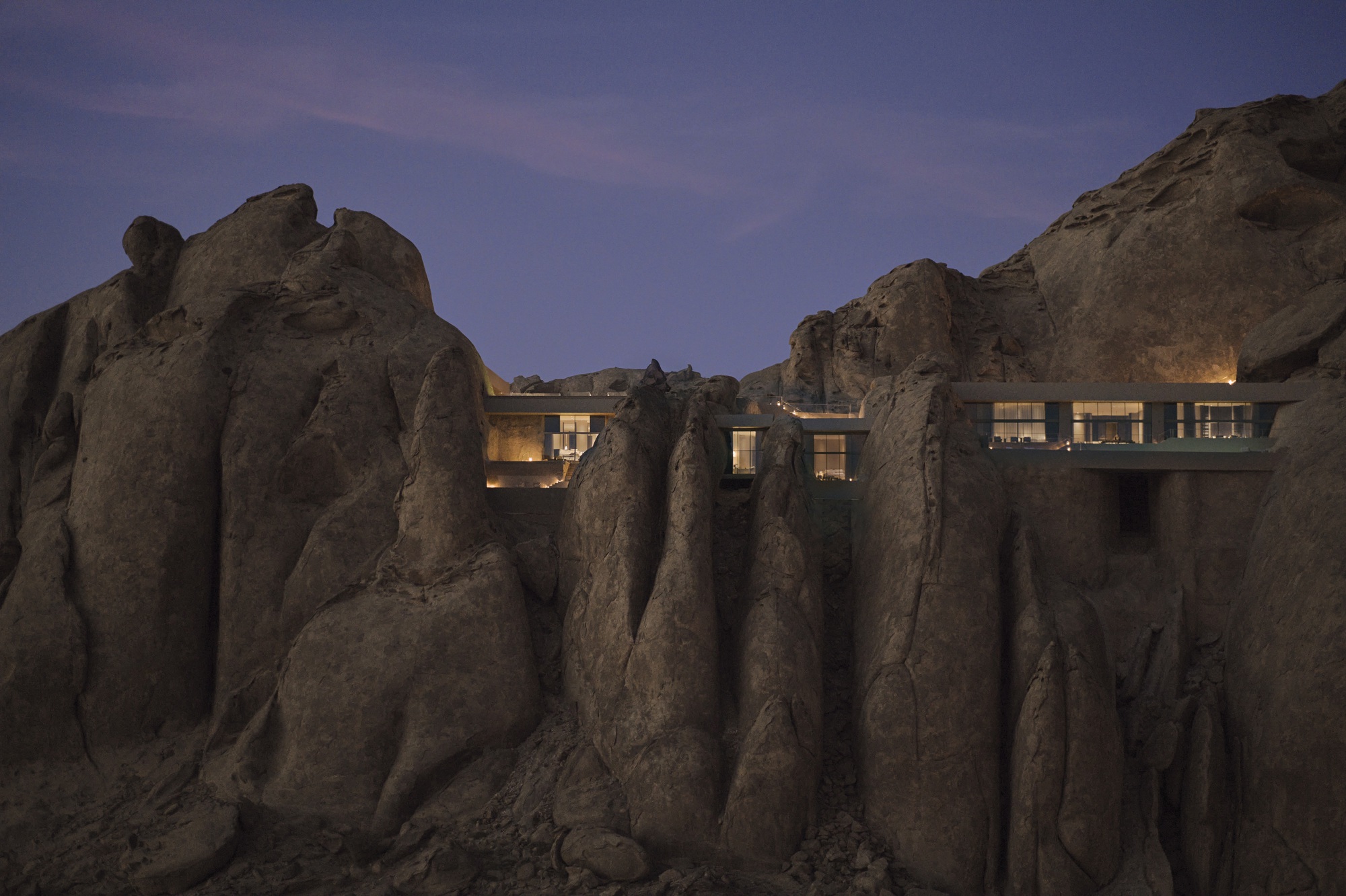 Illuminated glazed openings visible between vertical rock formations at twilight
