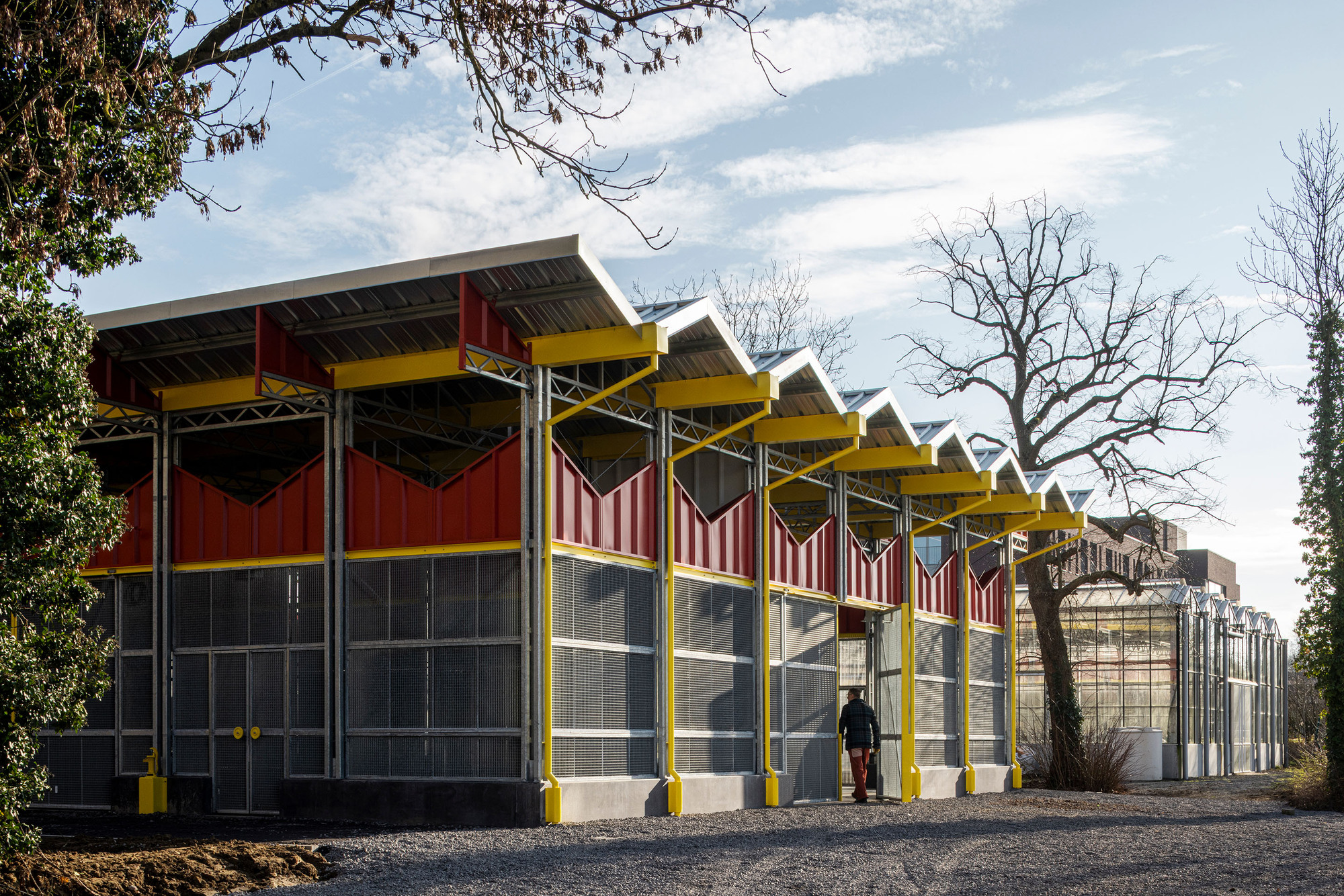 Side view of the sawtooth roof structure with bare winter trees and a person walking on the gravel path