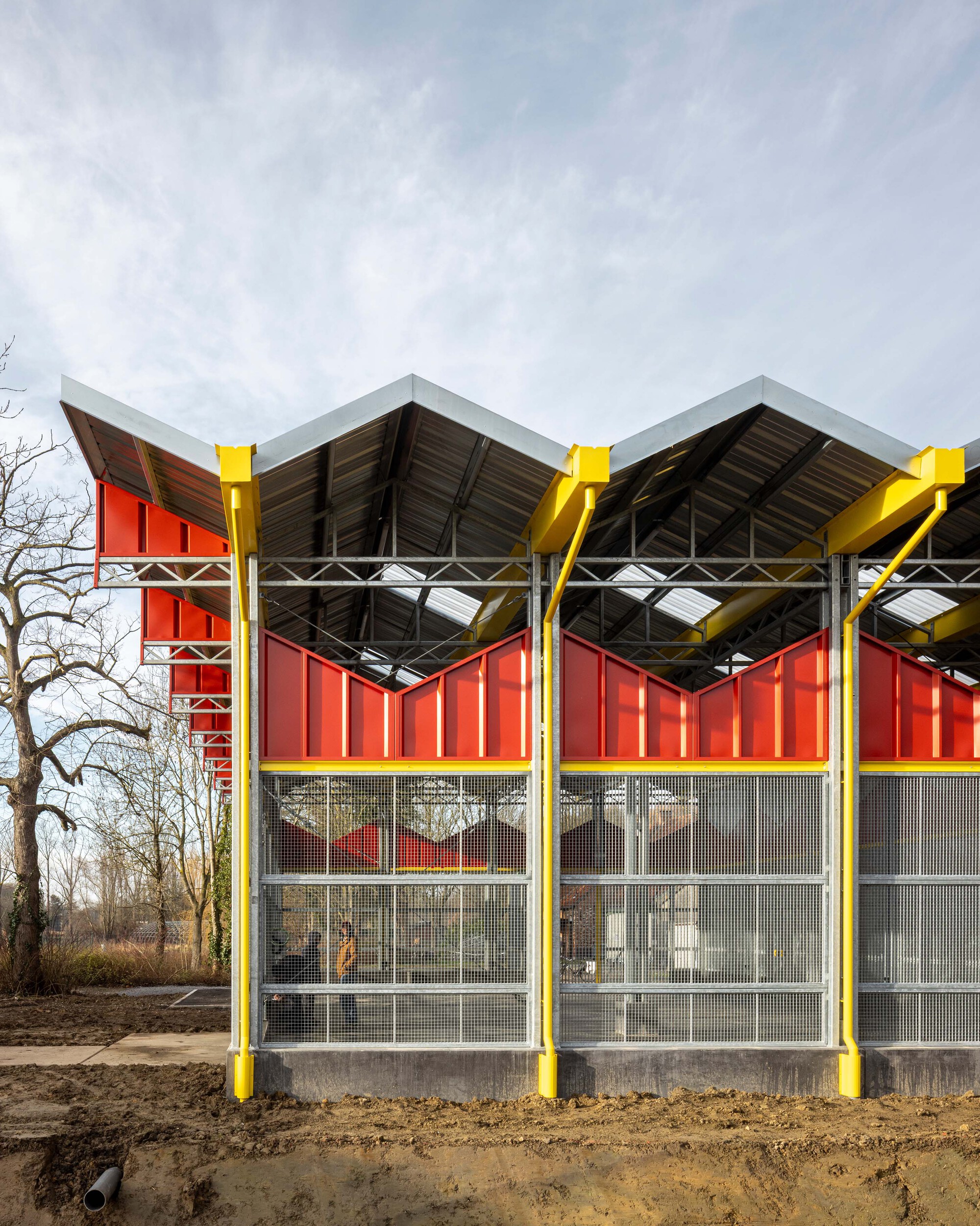 Facade with yellow steel columns, red corrugated panels, and mesh glazing beneath a sawtooth roof on bare ground