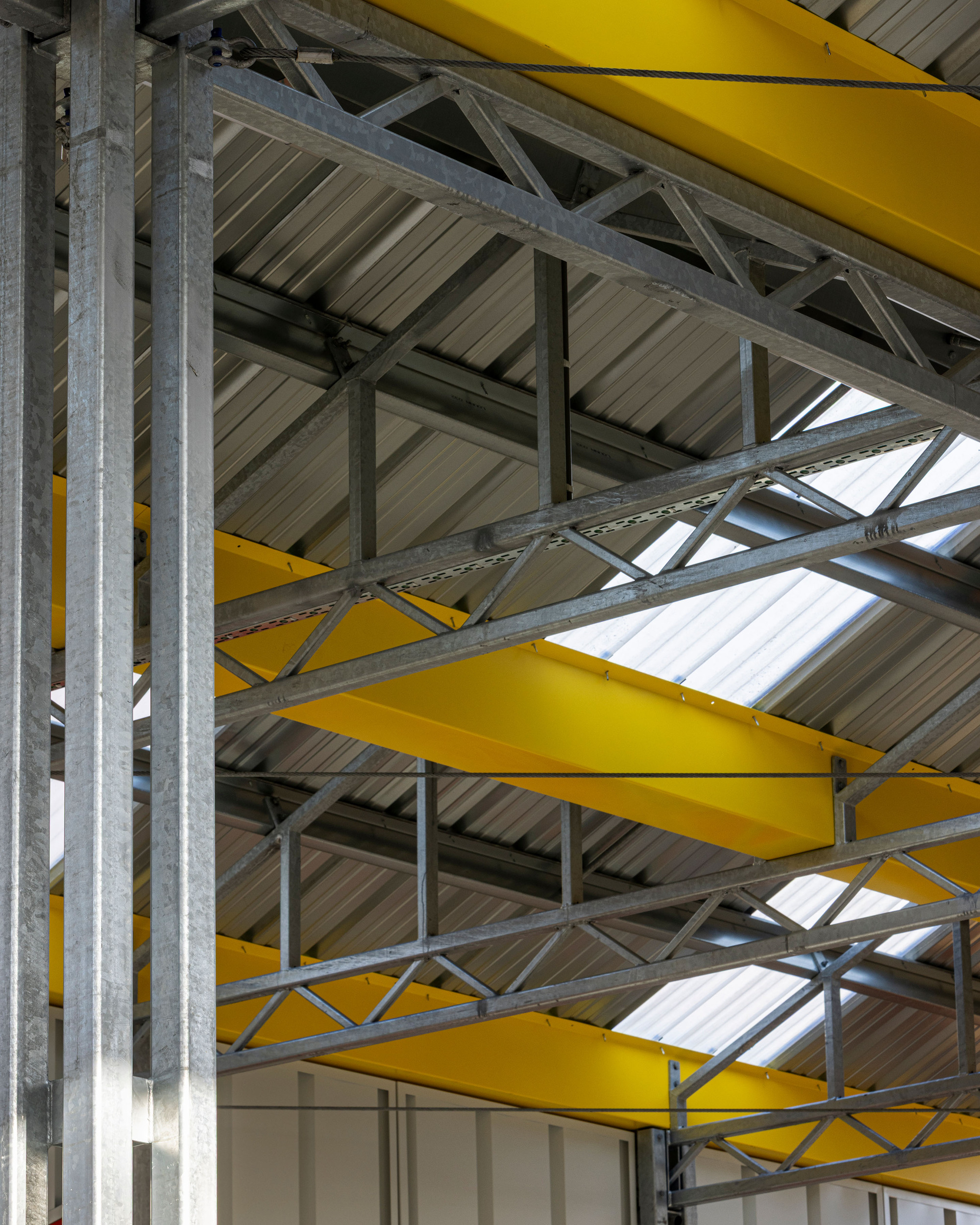 Upward view of yellow steel beams, diagonal trusswork, and corrugated metal ceiling with strip skylights
