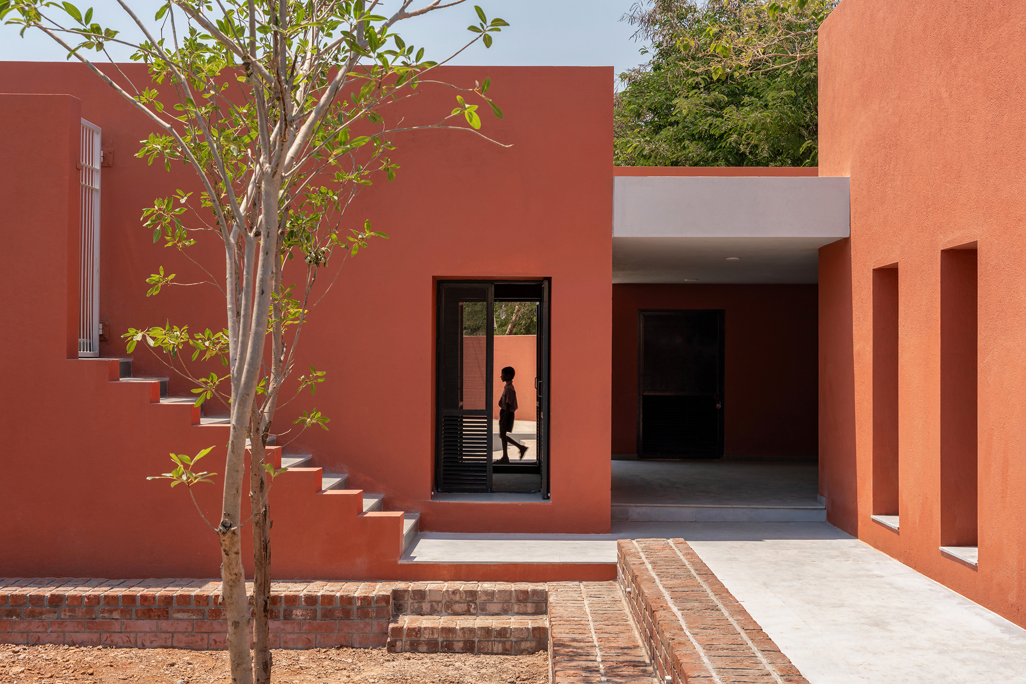 Outdoor staircase against the terracotta wall with a young tree and a figure in the doorway