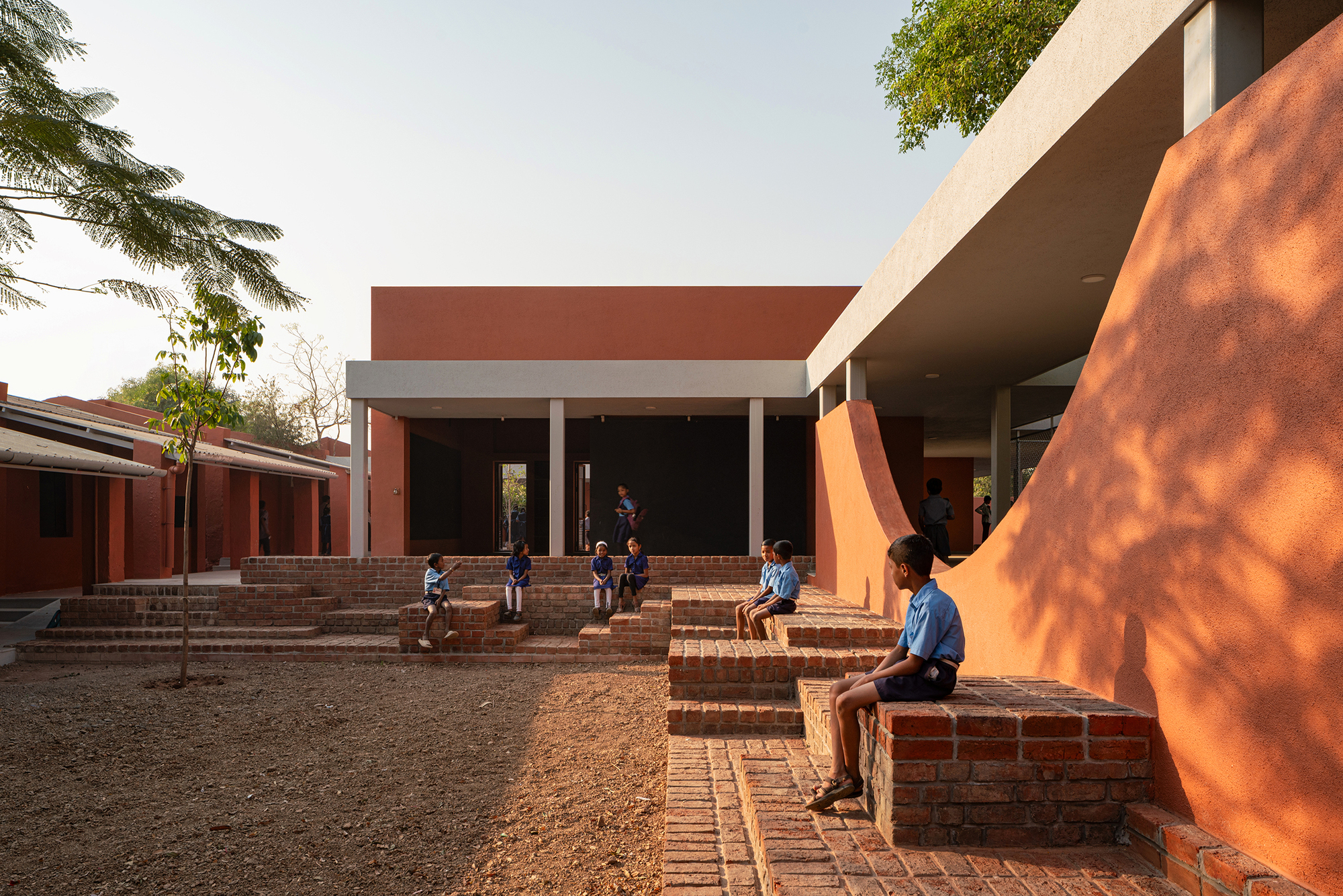 Courtyard with tiered brick seating and children gathering beneath the cantilevered concrete roof in late afternoon sun