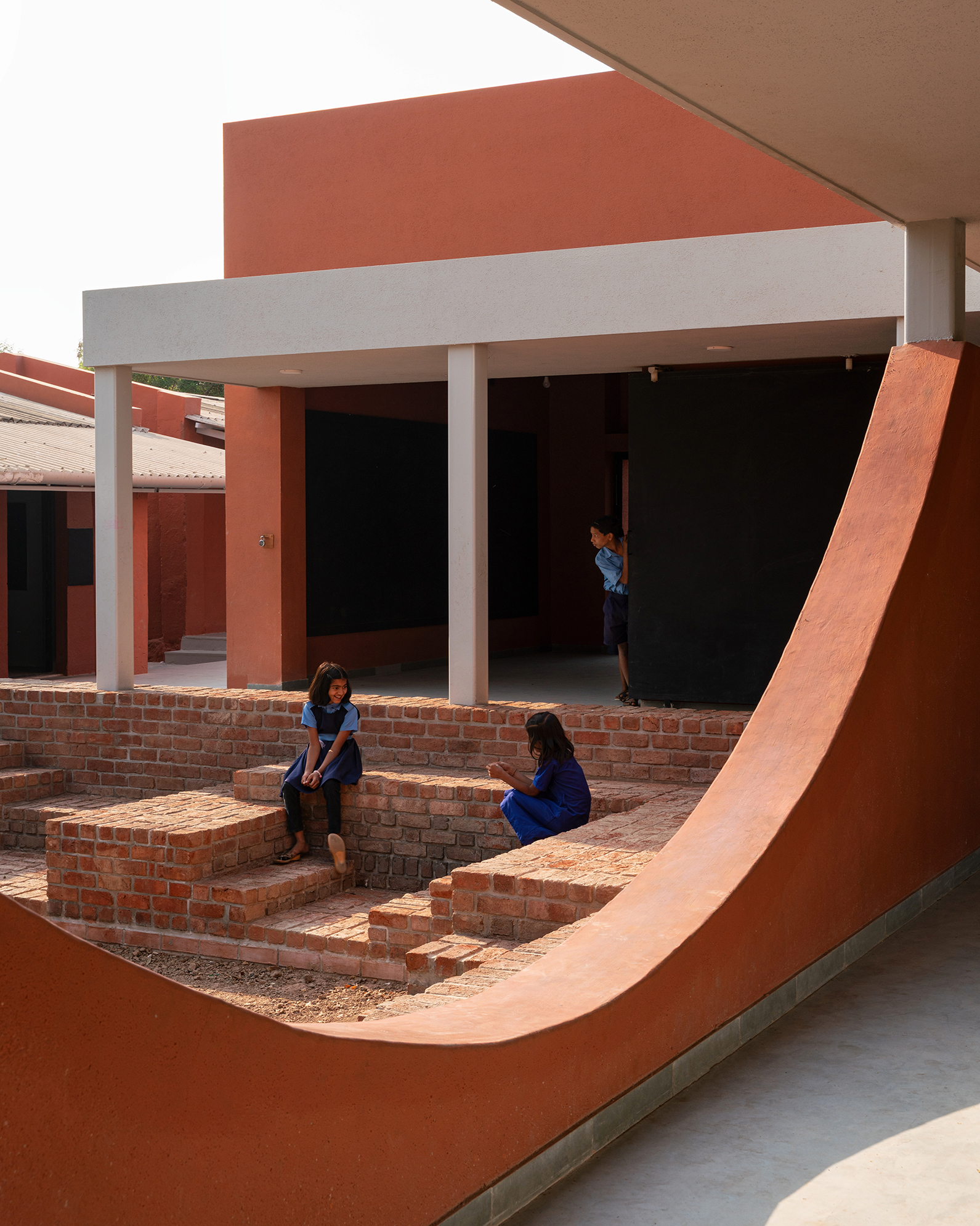View through the curved terracotta frame toward brick seating steps where children sit in the sheltered courtyard