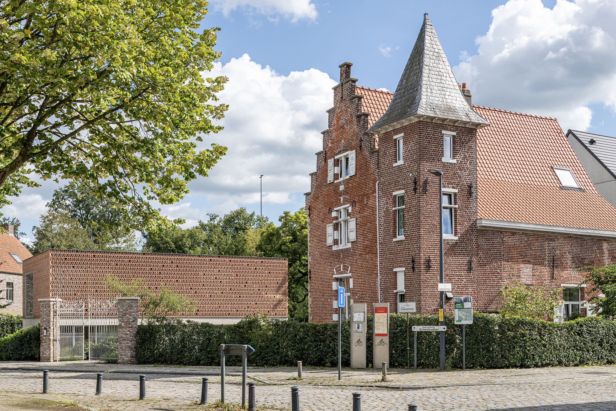 Low perforated brick addition adjacent to historic turret building with cobbled street and trees