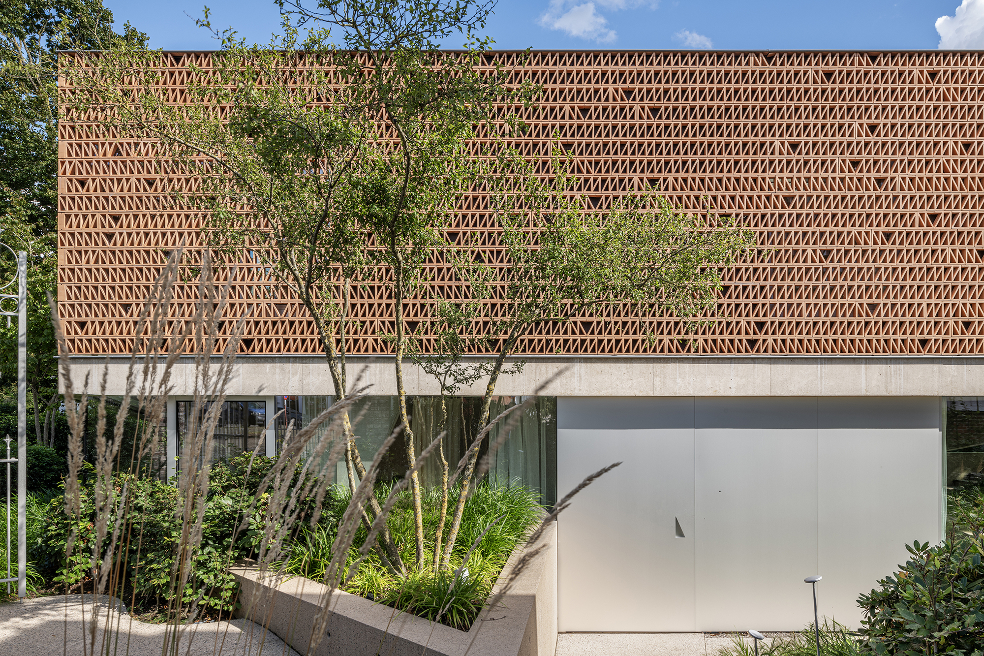 Perforated brick facade with young trees and planted beds in summer sunlight