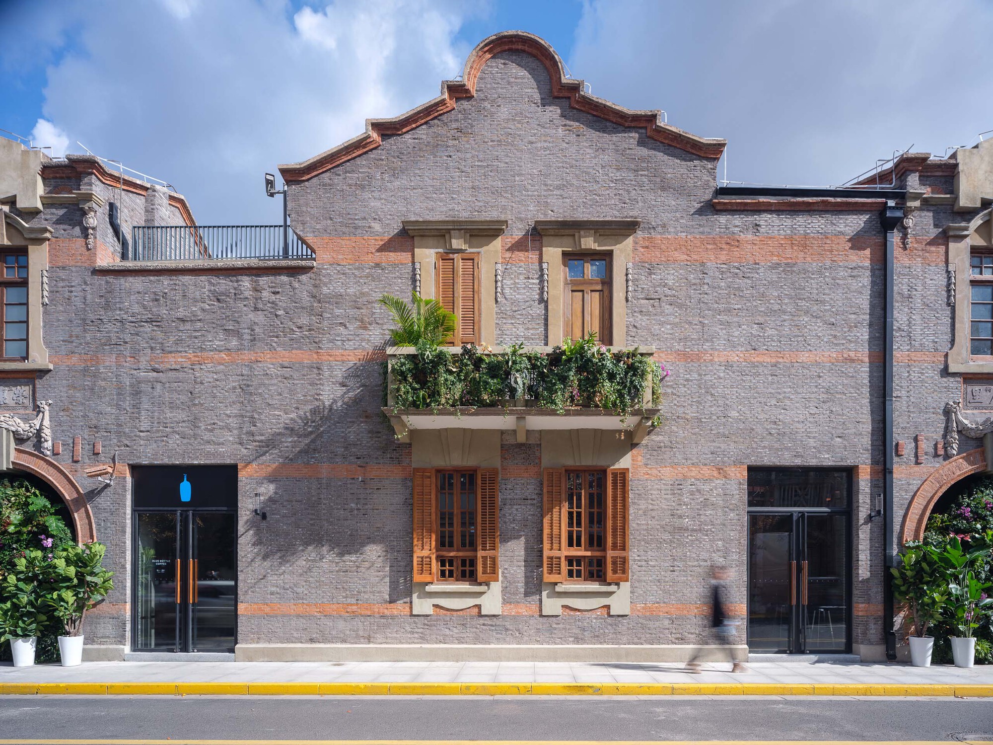 Street facade with painted brick, curved parapet, timber doors and balcony planters under scattered clouds