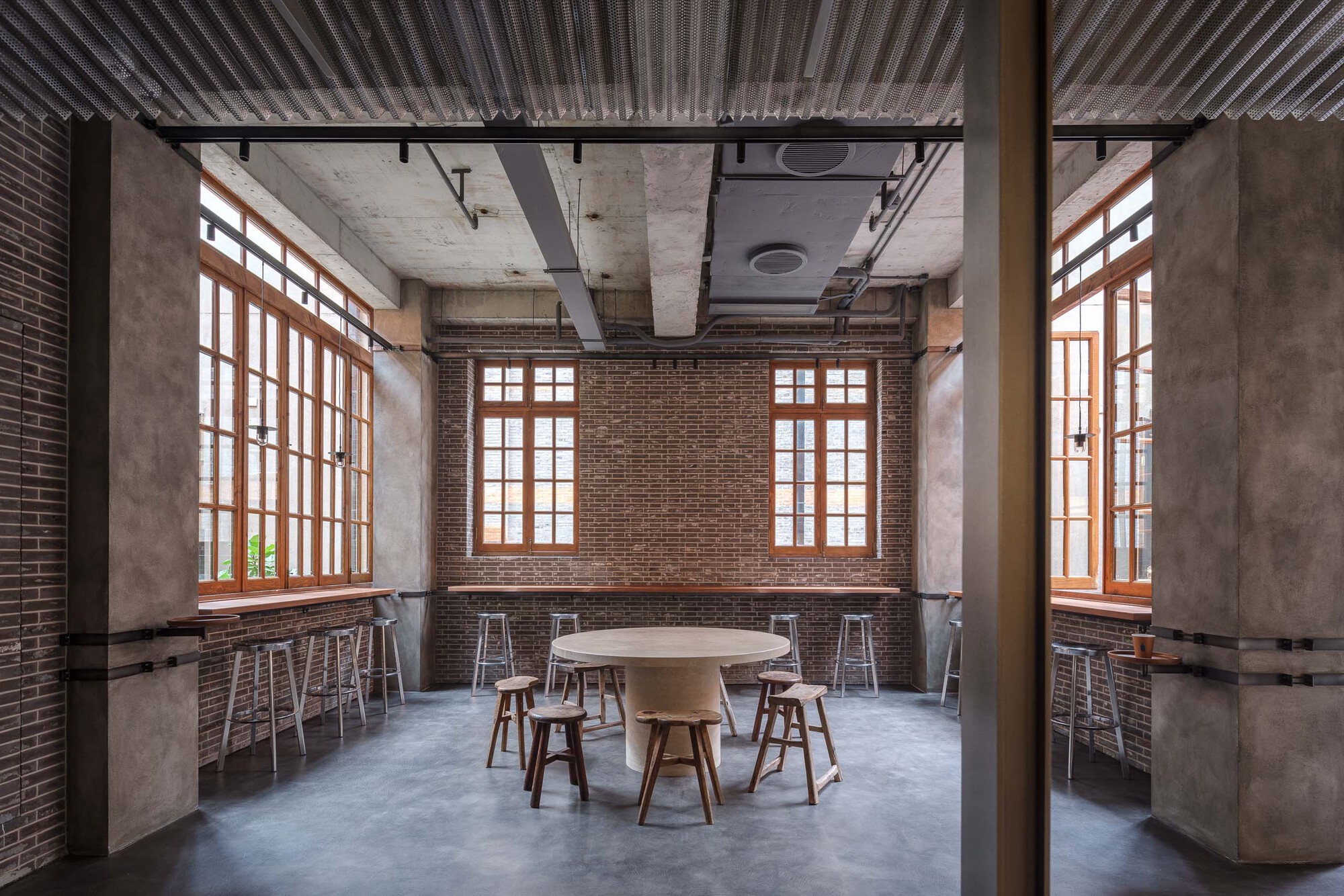 Seating area with timber-framed windows, exposed brick walls, round table and stools beneath corrugated metal ceiling