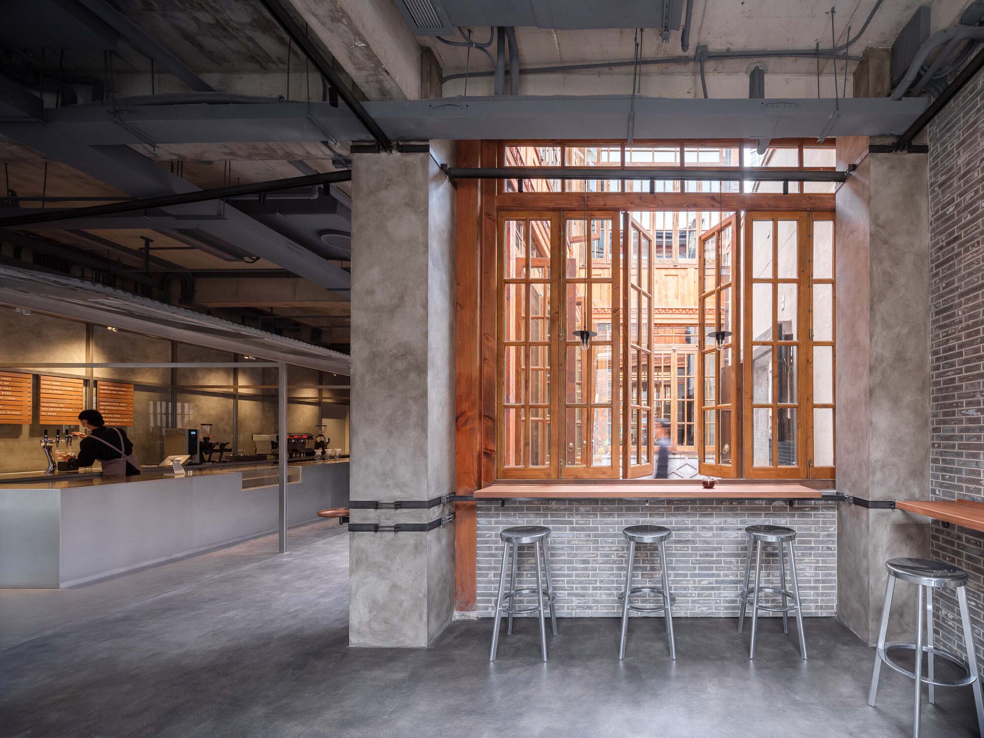 Brick bar counter beneath timber-framed windows with metal stools and a barista working in background
