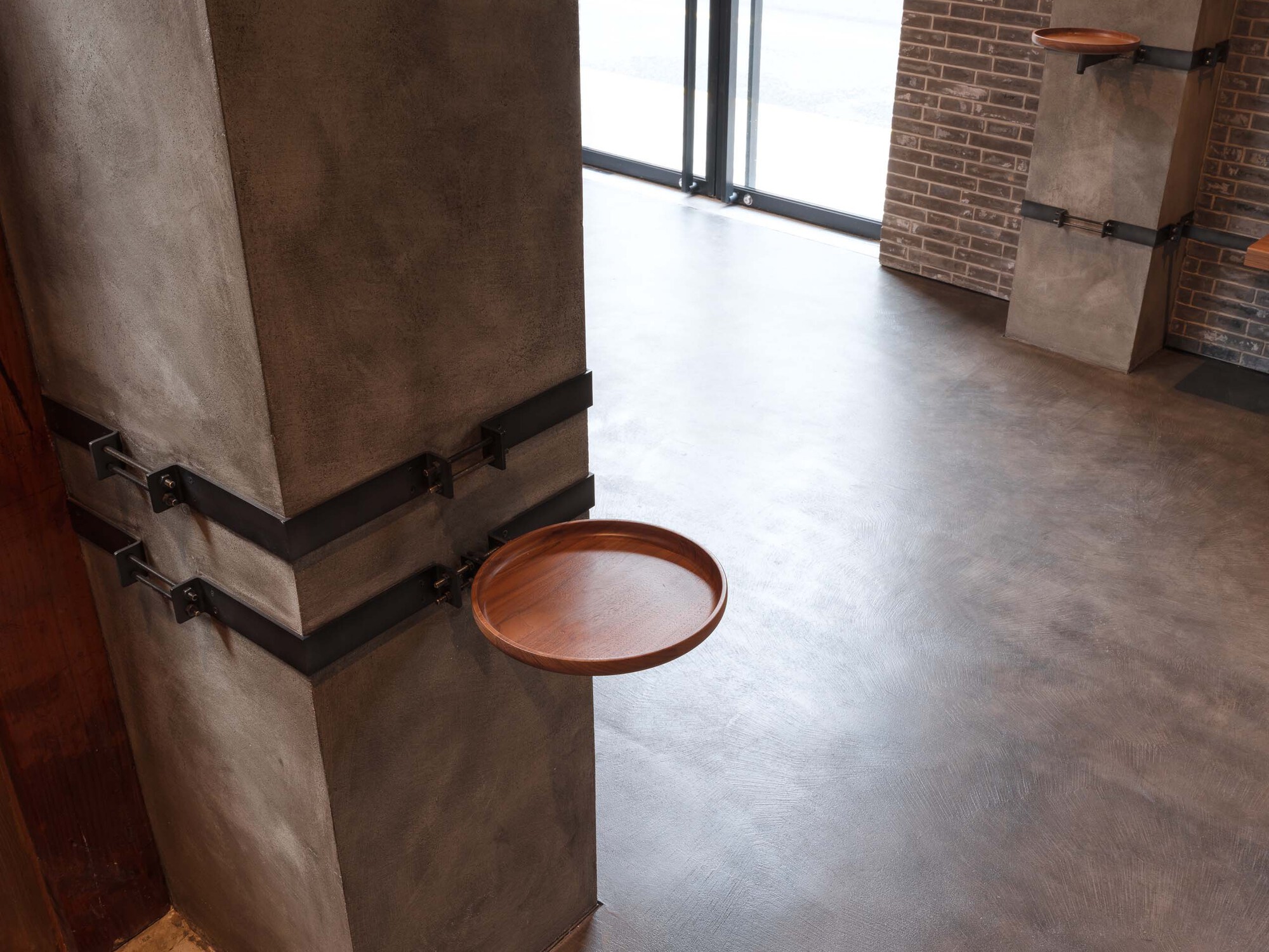 Concrete column wrapped with black steel bands and wooden tray shelf overlooking polished concrete floor