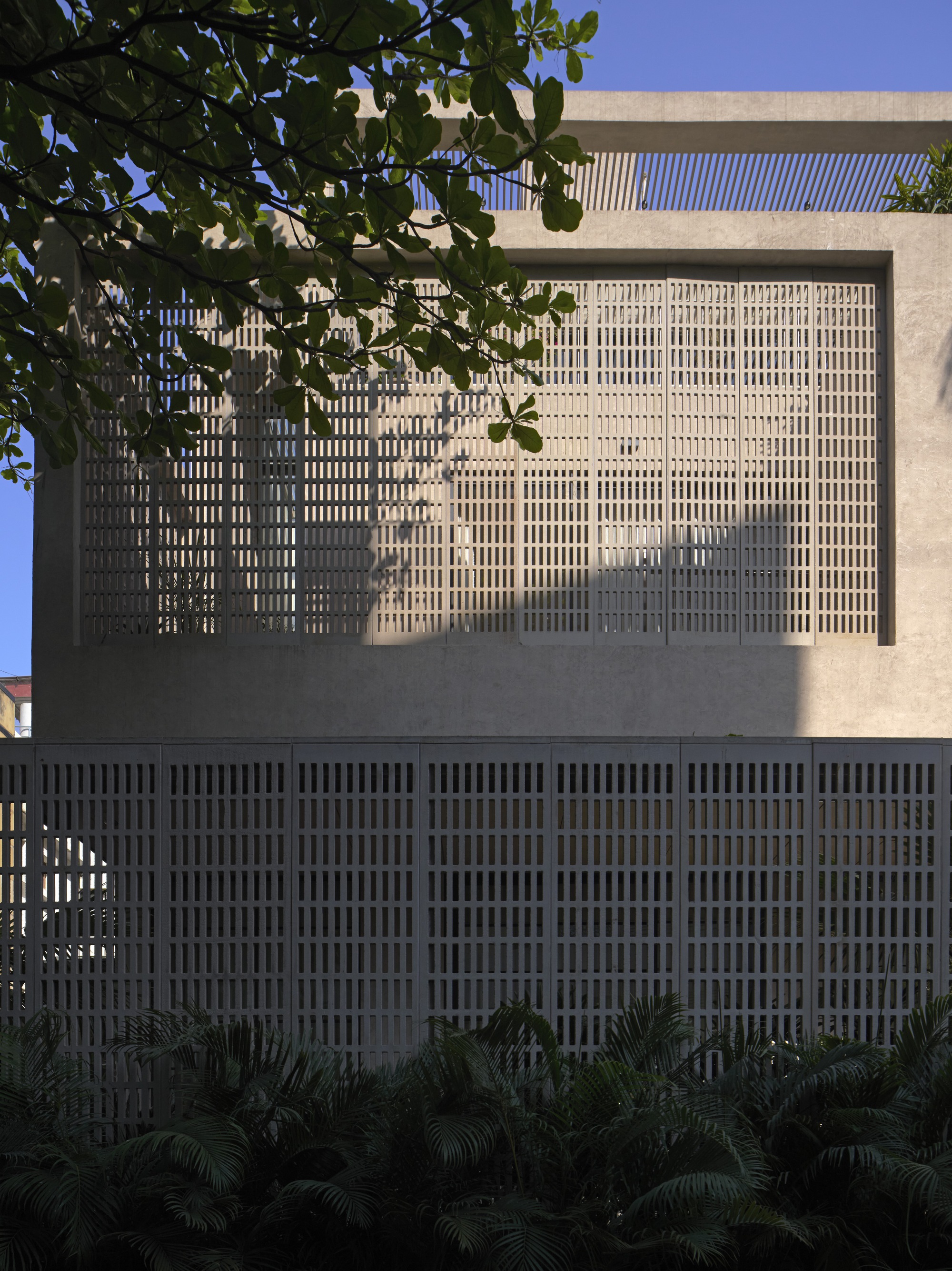Perforated brick screen facade casting dappled shadows from overhanging foliage onto the concrete frame