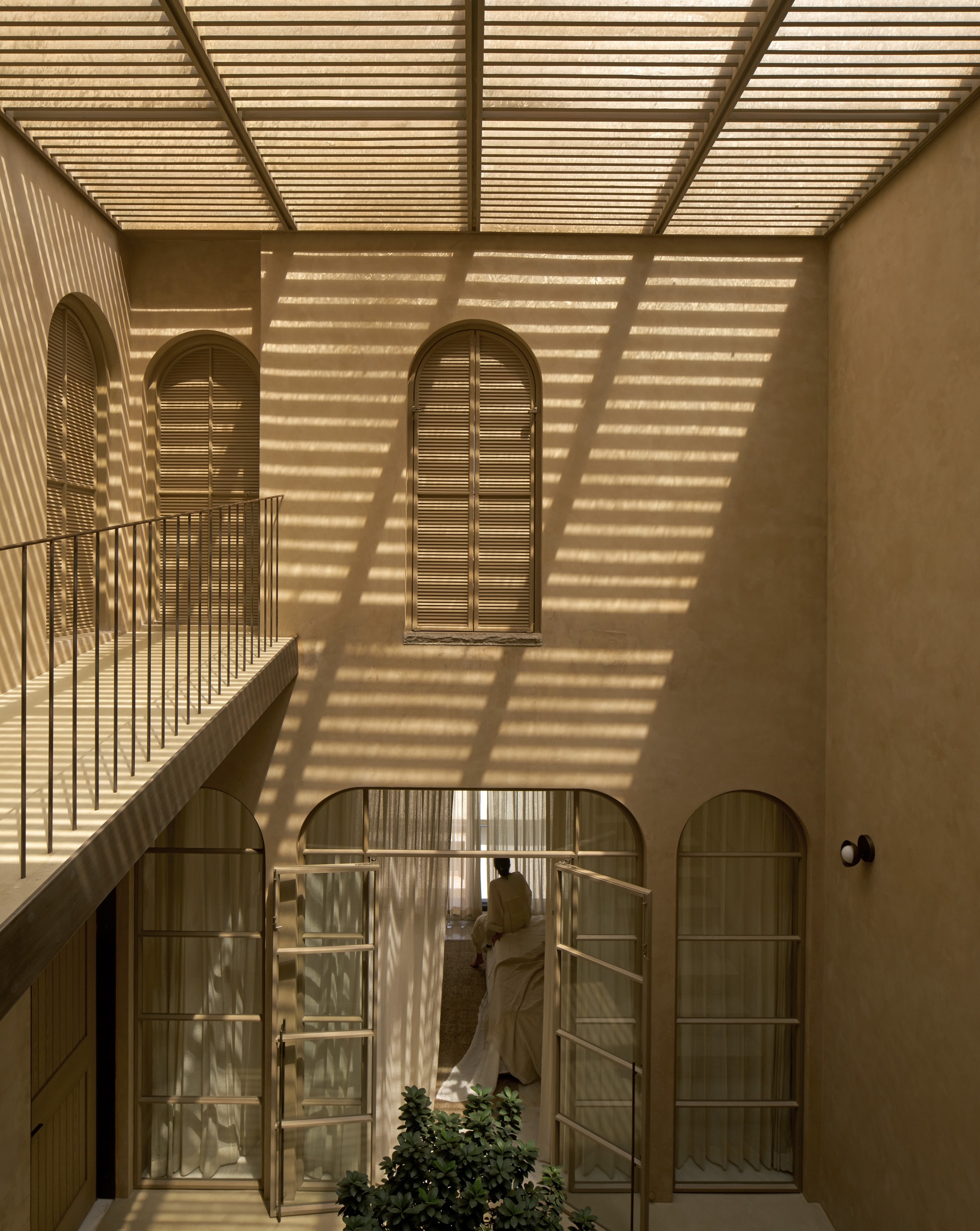 Interior courtyard with arched openings and timber louvered skylight casting striped shadows across plaster walls