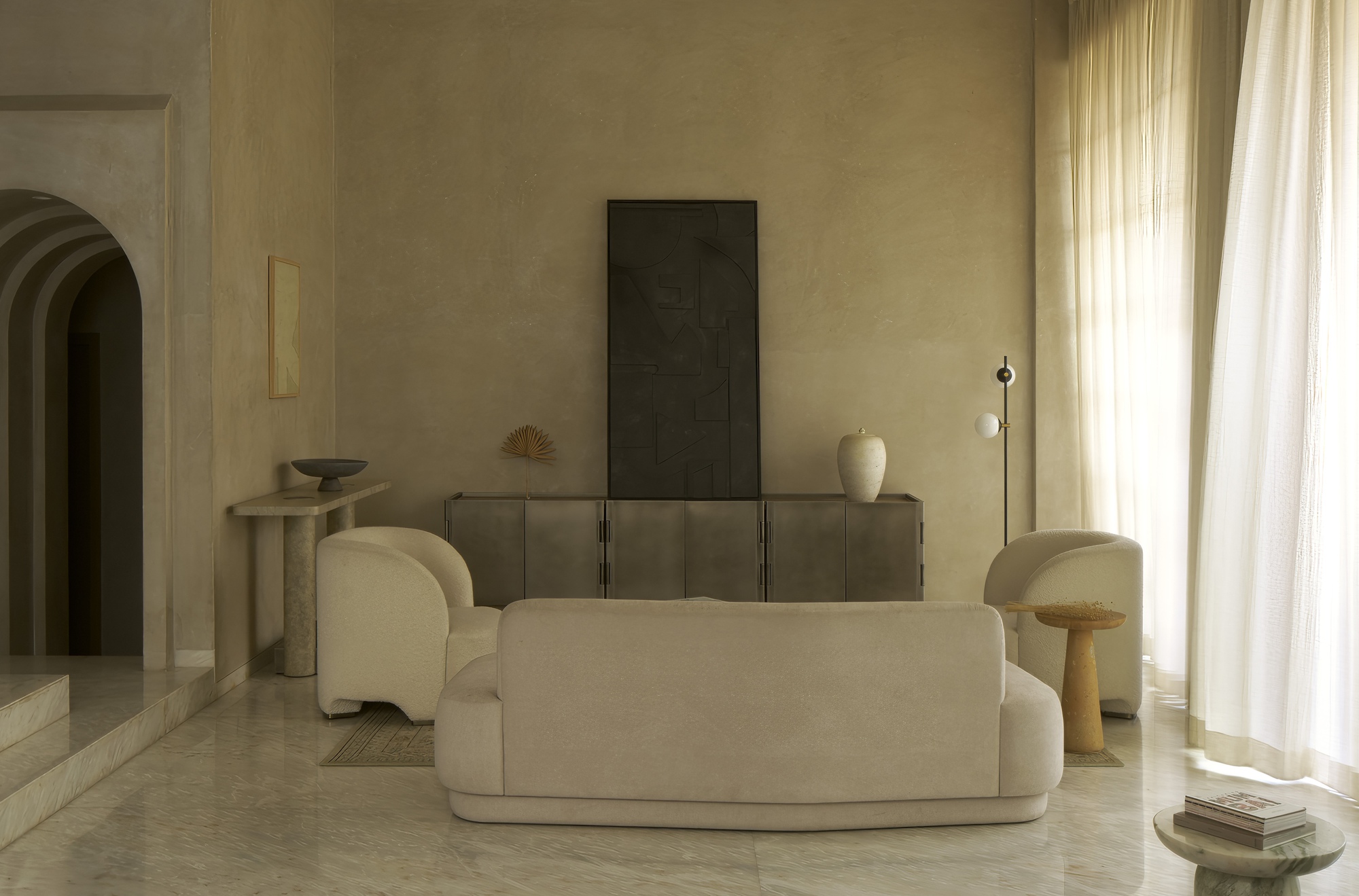 Living room with rounded upholstered furniture, timber credenza, and sheer curtains filtering daylight