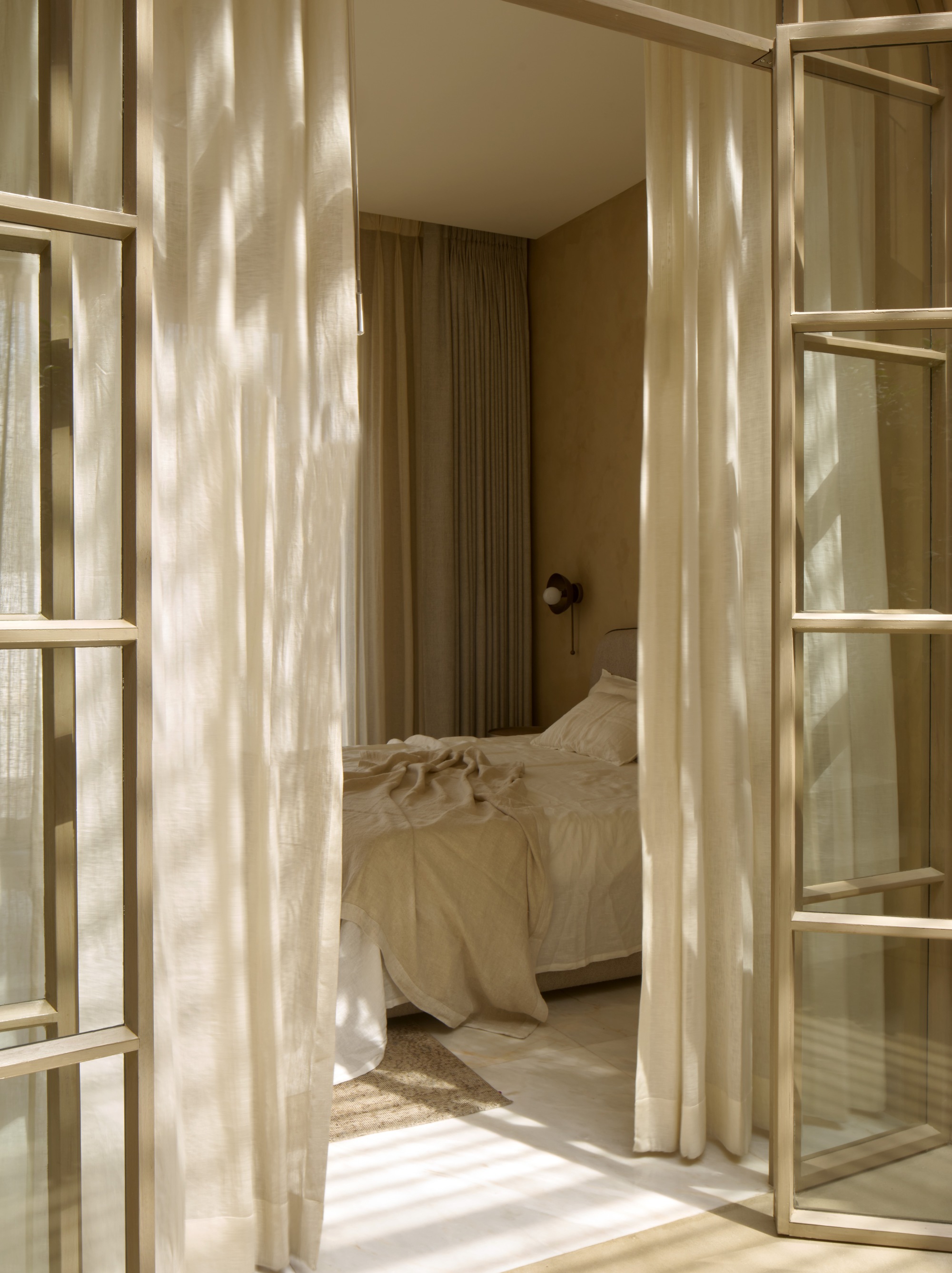 Bedroom view through open timber doors with sunlight casting shadows on rumpled linen bedding