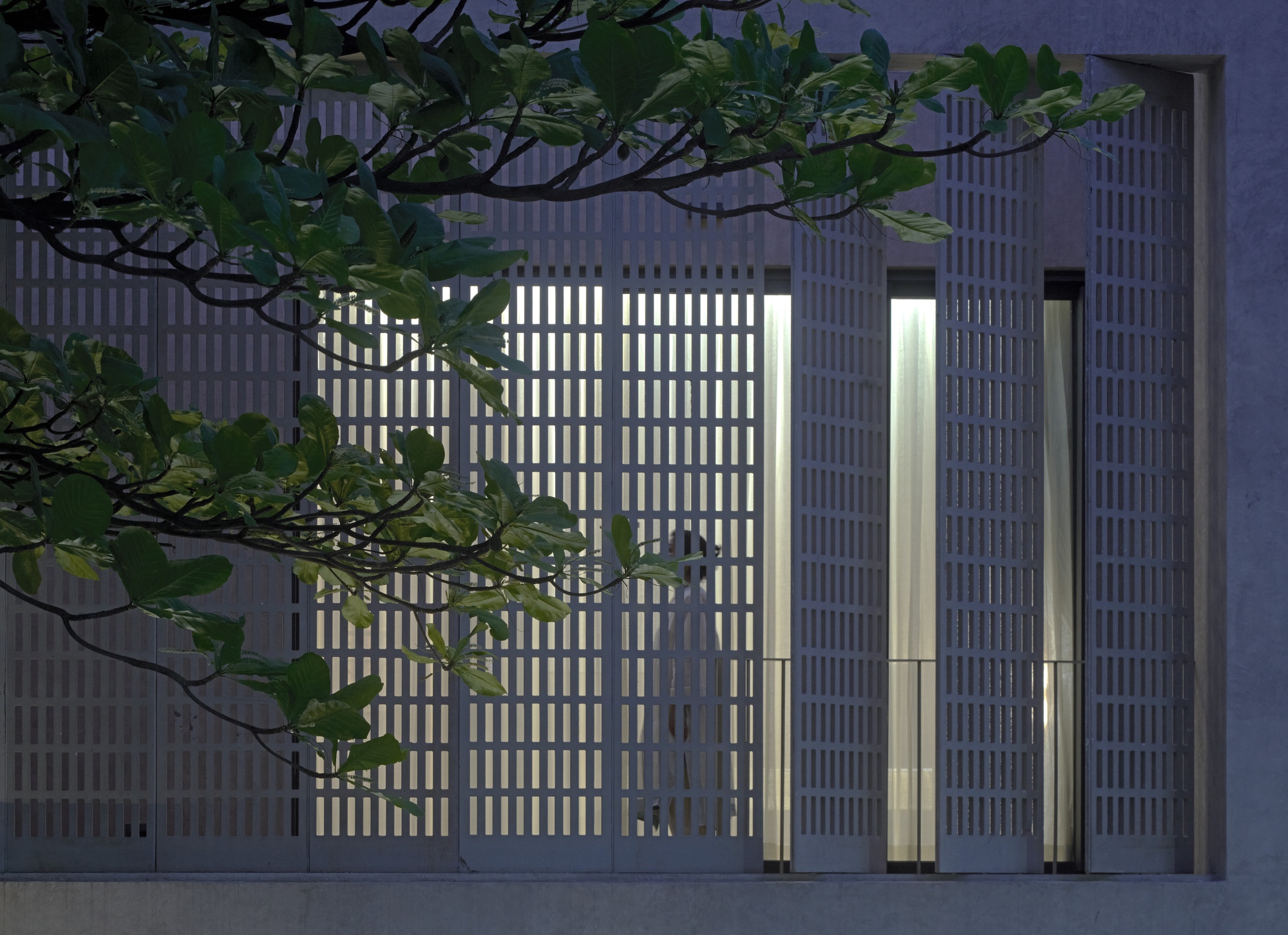 Illuminated vertical-slat facade at dusk with tree branches crossing the perforated concrete screen