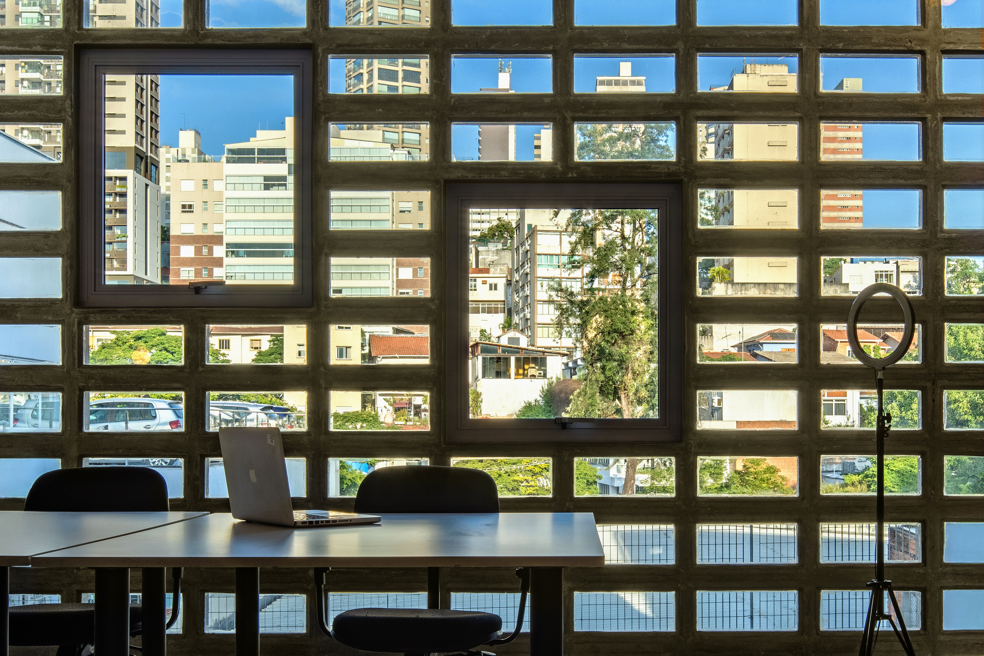 Interior view through concrete grid screen framing a table with laptop overlooking neighboring buildings and trees