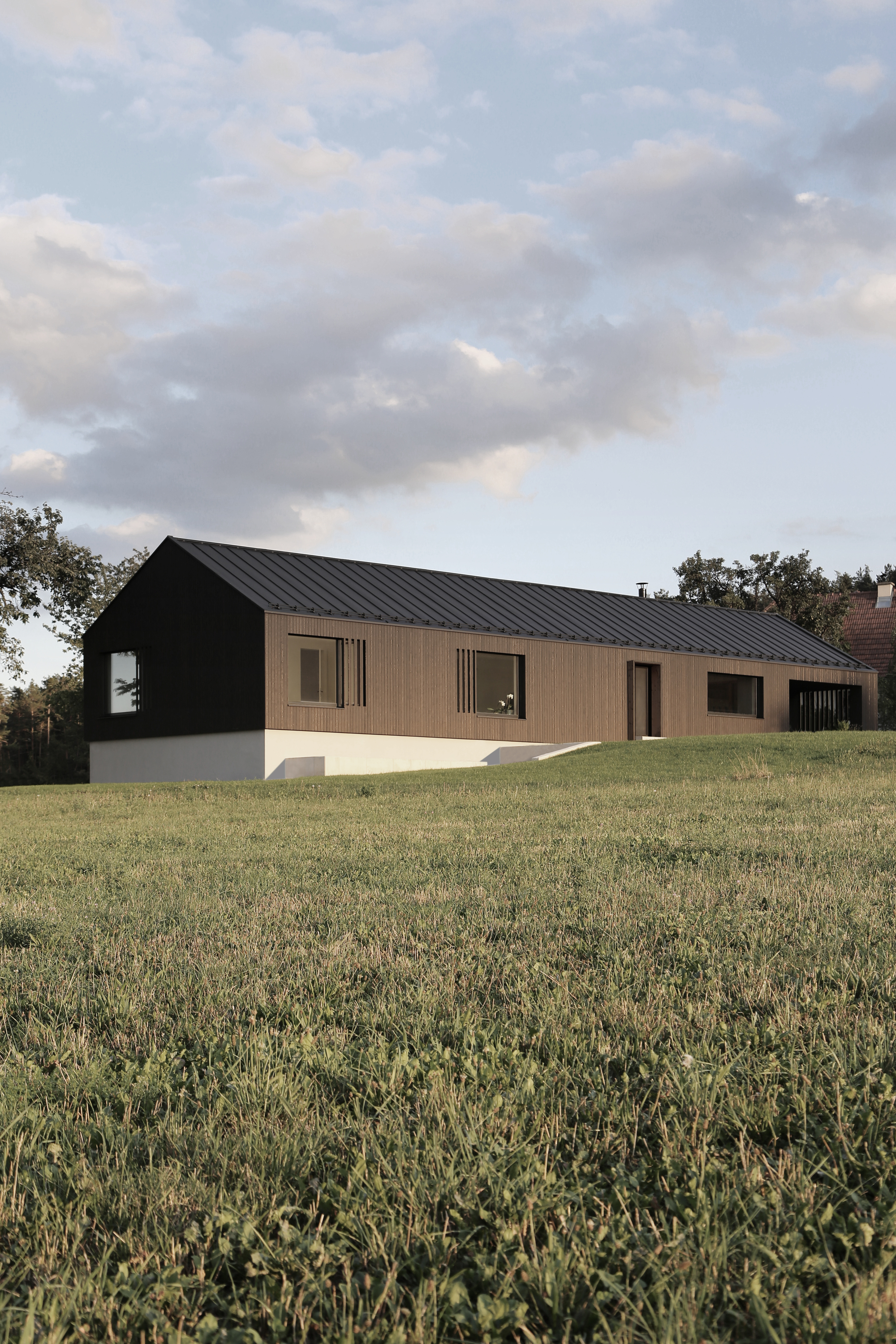 Long elevation showing the dark metal gable roof extending across black and timber-clad facades at dusk