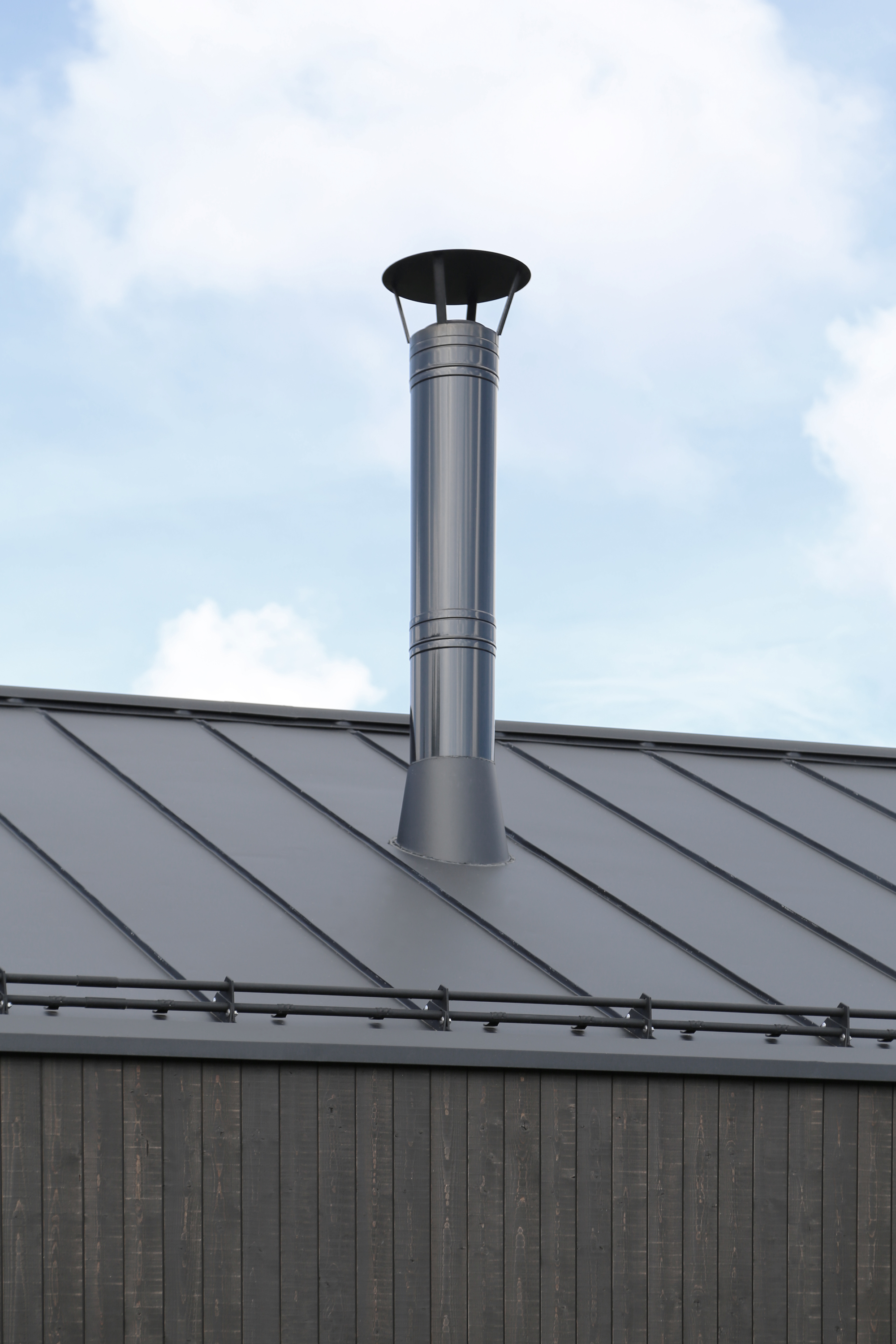 Stainless steel chimney pipe rising from a standing seam metal roof against a cloudy sky