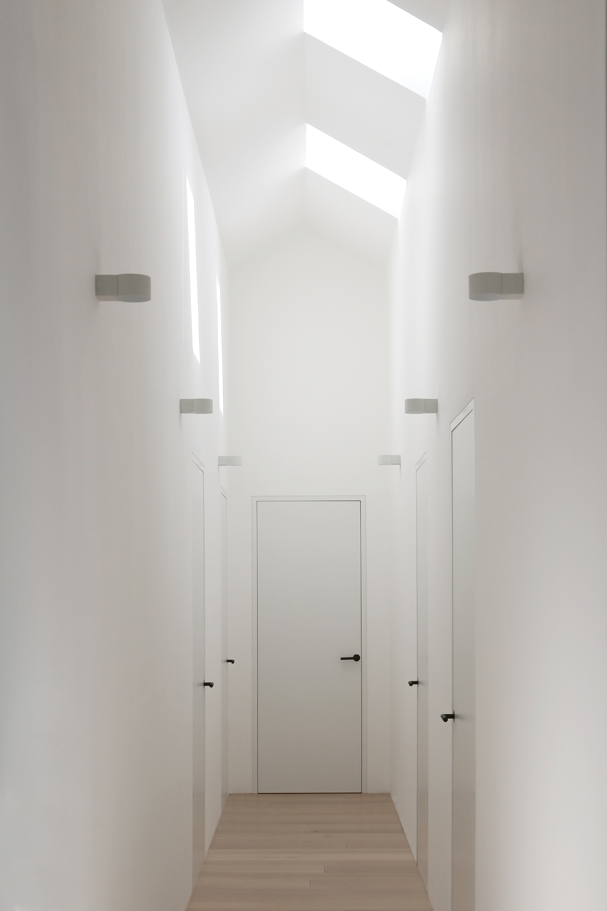 Narrow white hallway with concealed wall sconces and multiple skylights casting bright geometric shadows